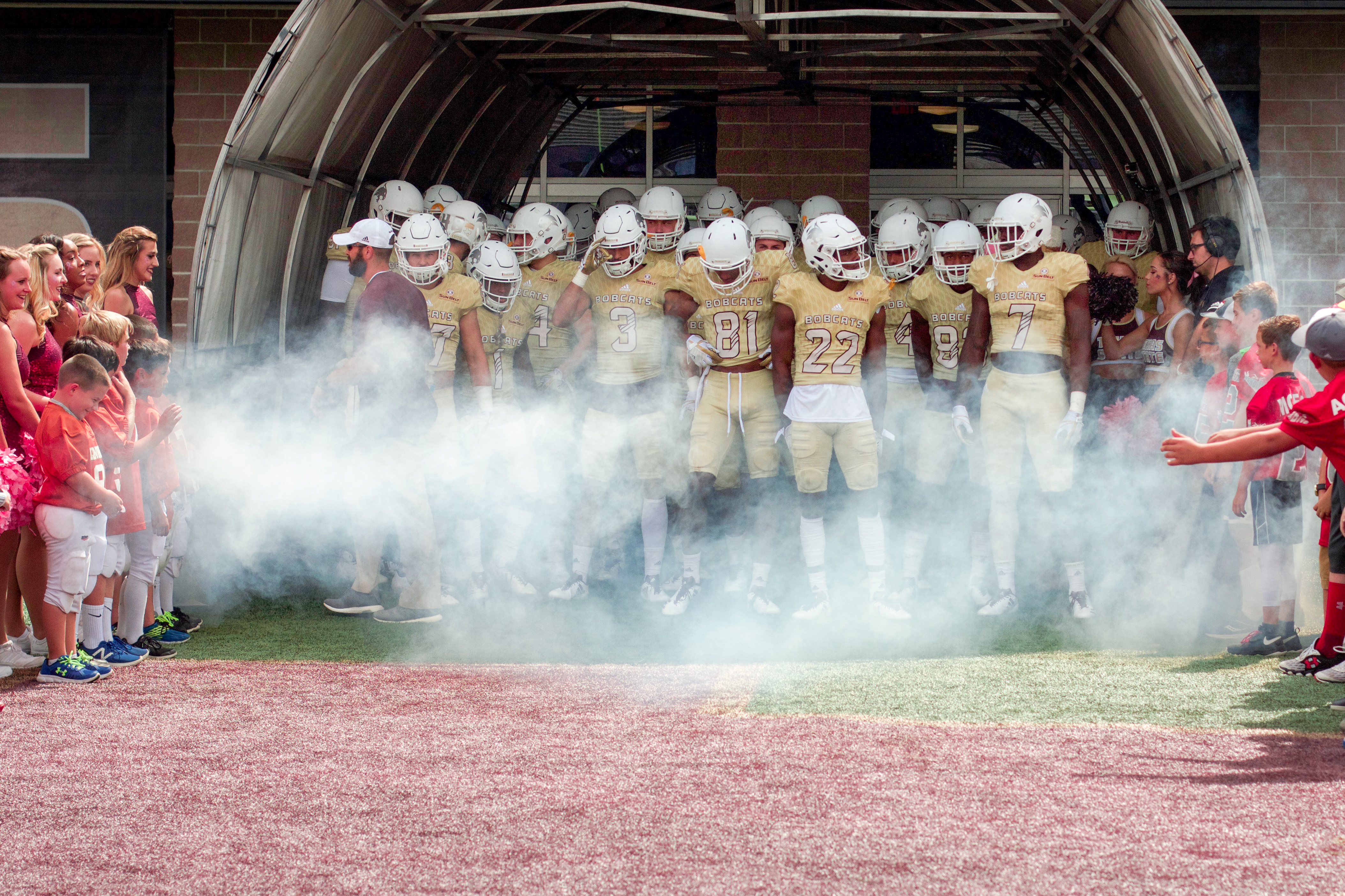In their bright golden jerseys, the Texas State Bobcat Football team prepares to rush out of a smoke-filled tunnel to an enthusiastic home crowd. Some players look at each other, others look down, but all have rigid bodies from the tense of the situation, waiting to pour their exuberance onto the field of play.