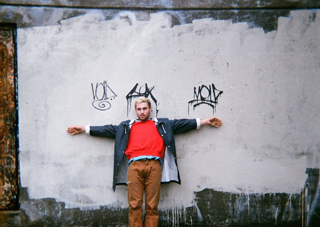 Yoke Lore standing in front of a building wall with graffiti tags above him
