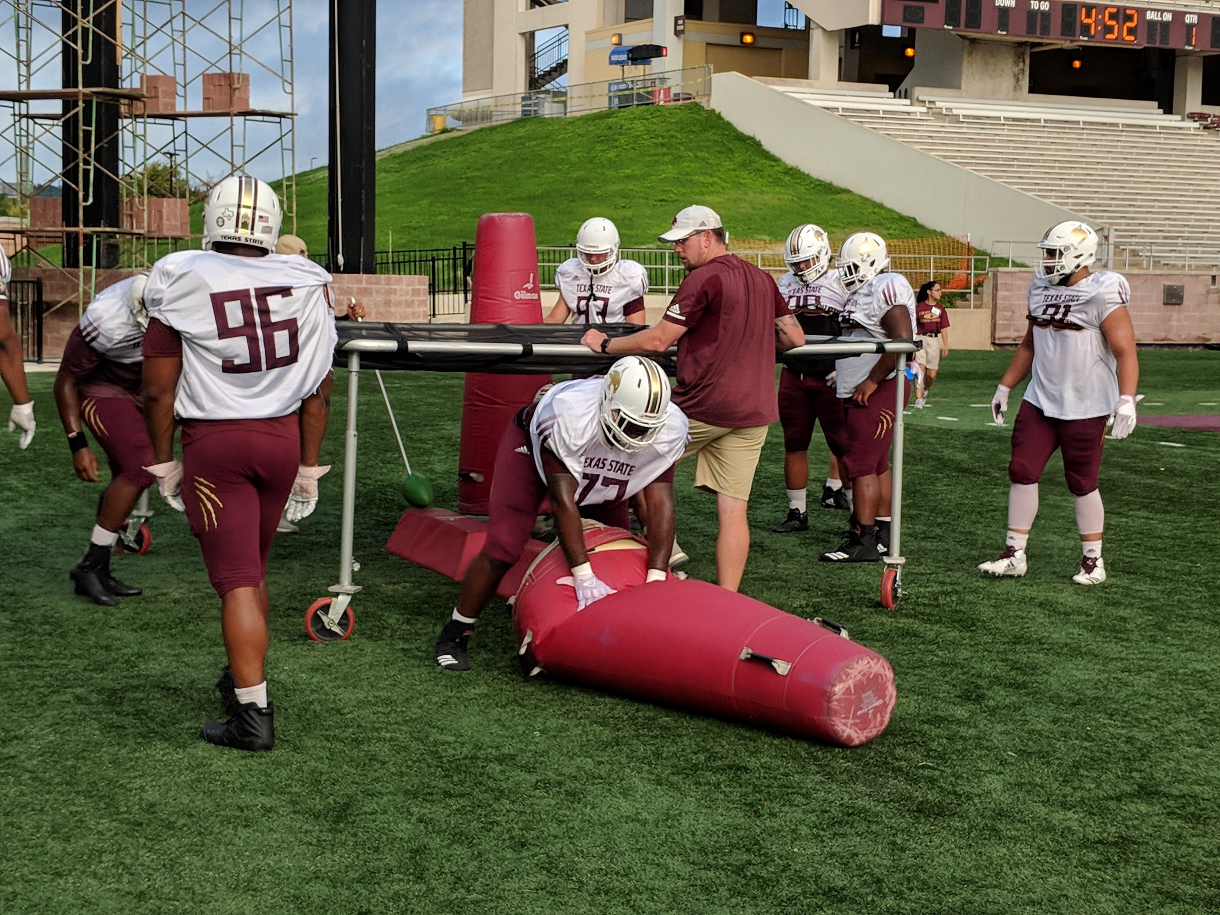 On a cloudy day in San Marcos, the defensive line, in white jerseys, practices scurrying under a netting to pop a practice dummy bag. Number 73 Jaquel Pierce is pictured finishing the tackling.