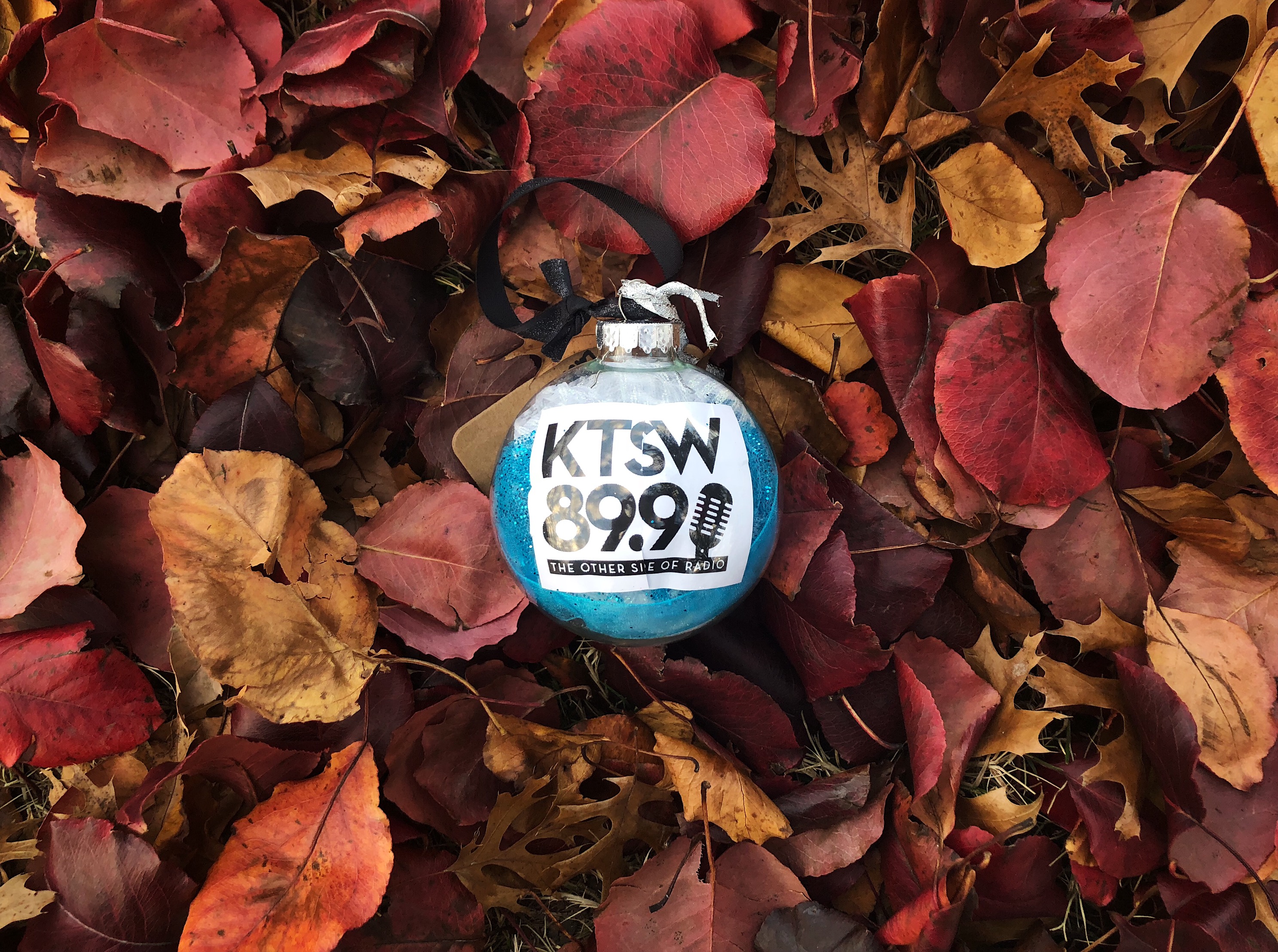 The photo features a clear ornament with sea blue tissue paper on the inside as well as a paper with the KTSW logo printed on it (KTSW 89.9: The Other Side of Radio). The ornament was photographed on a pile of fallen red, brown and yellow leaves.