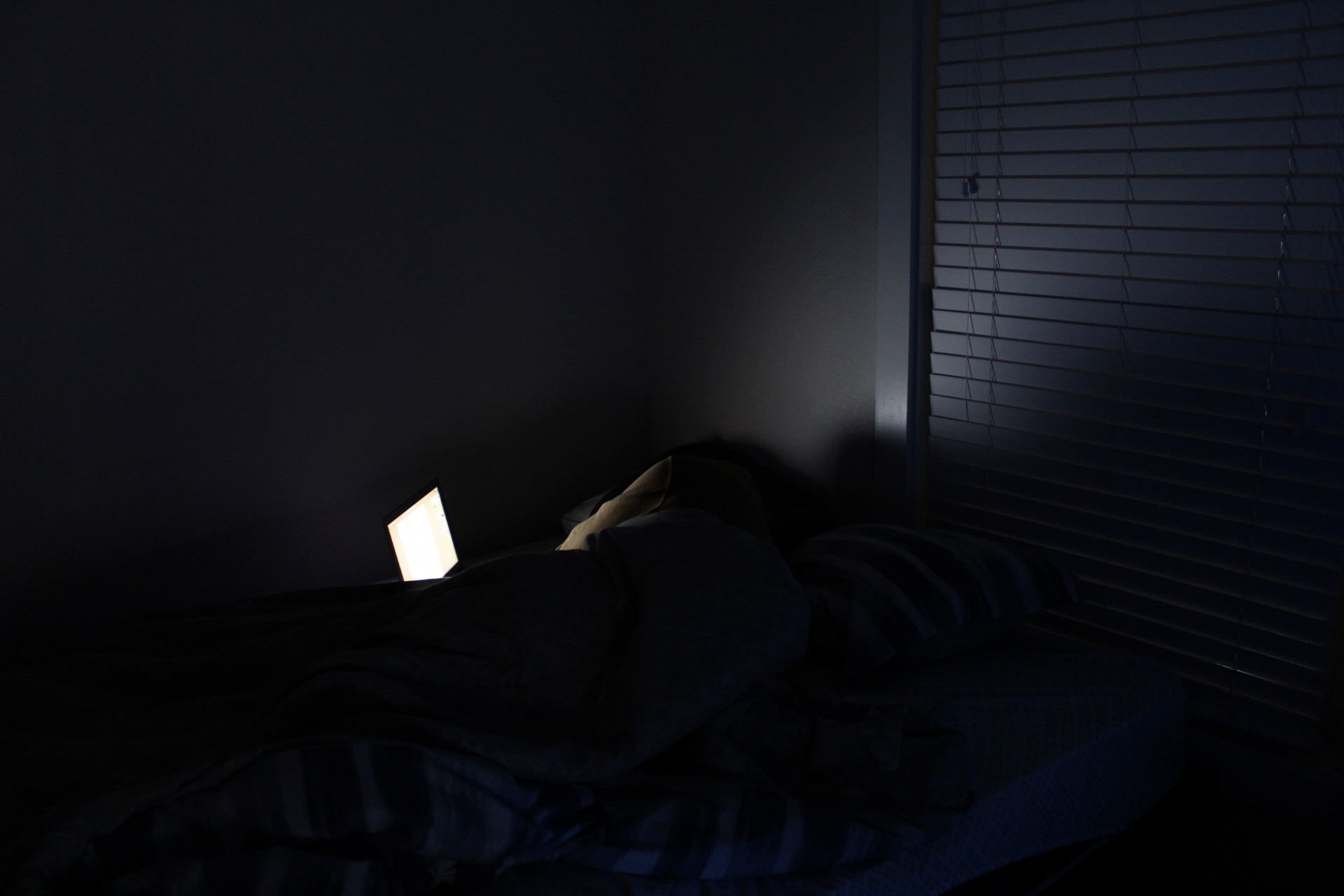 a guy in an orange jacket is laying in bed with a white light coming from a computer on the bed.