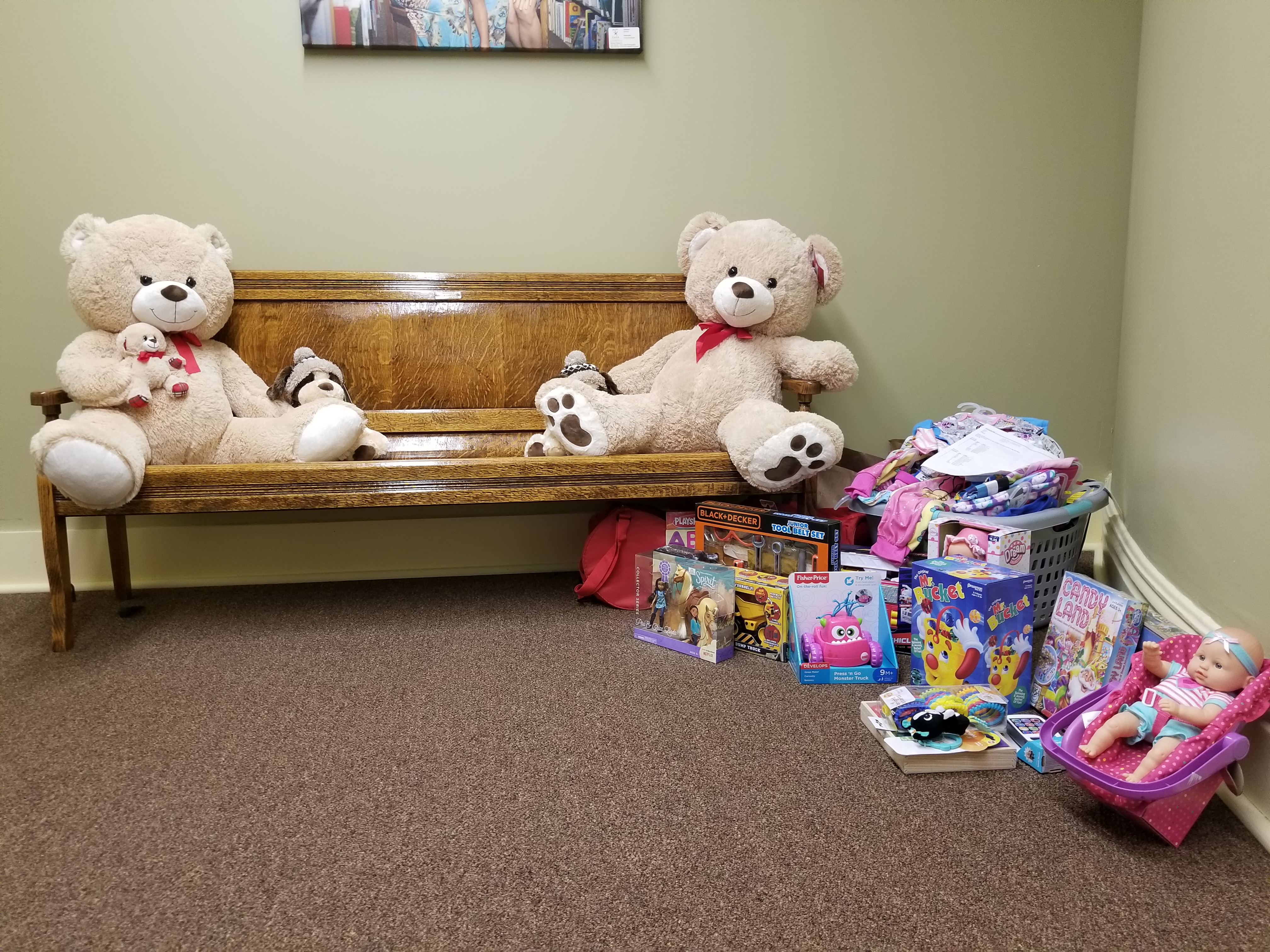 Two teddy bears sit opposite one another on a bench, to the right of the bench on the floor there is a pile of miscellaneous toys, and above the two bears a portrait can be barely be seen in the frame.