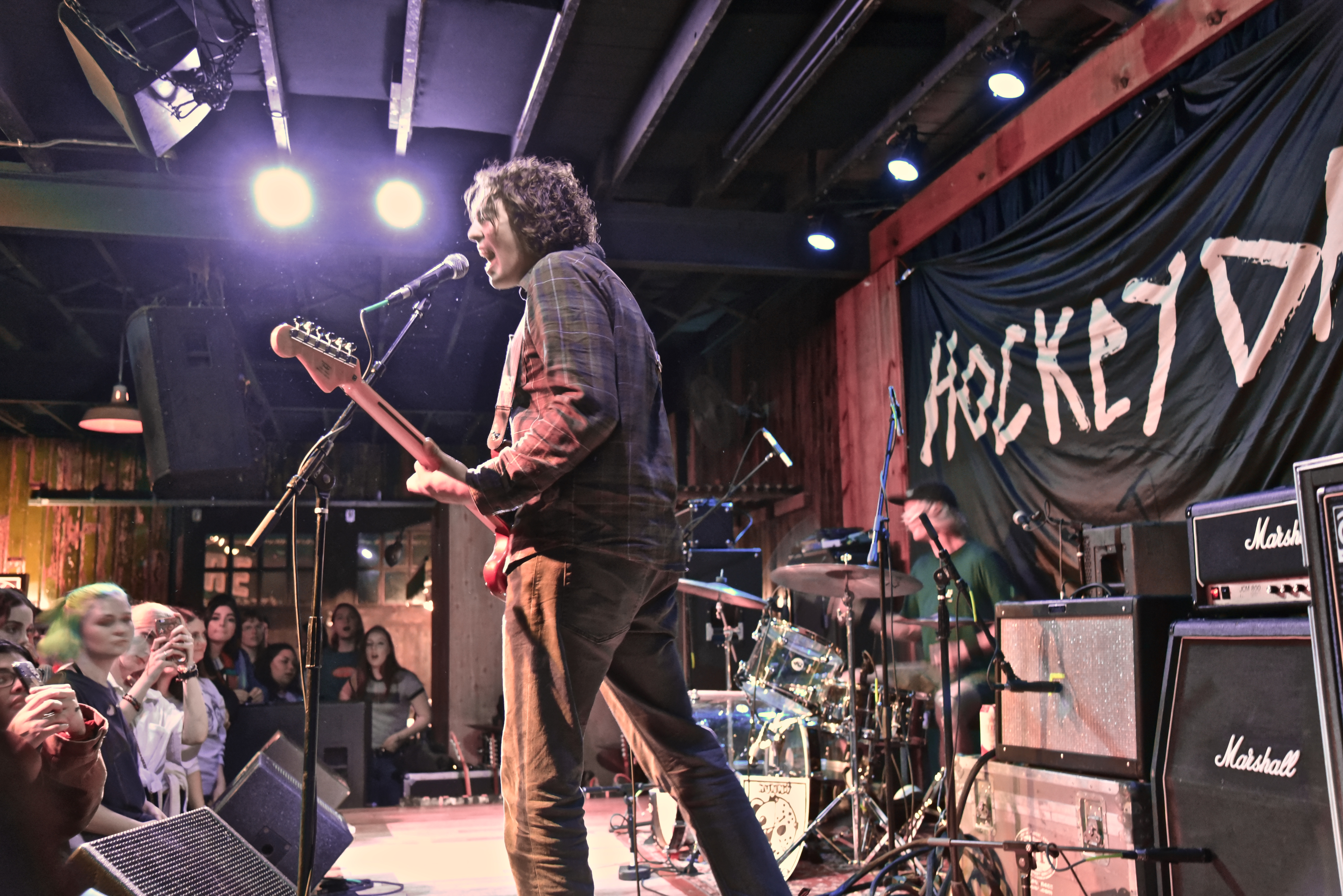 The lead singer and guitarist of Hockey Dad sings into the microphone while the drummer is in the background. Their “Hockey Dad” banner hangs in the back.
