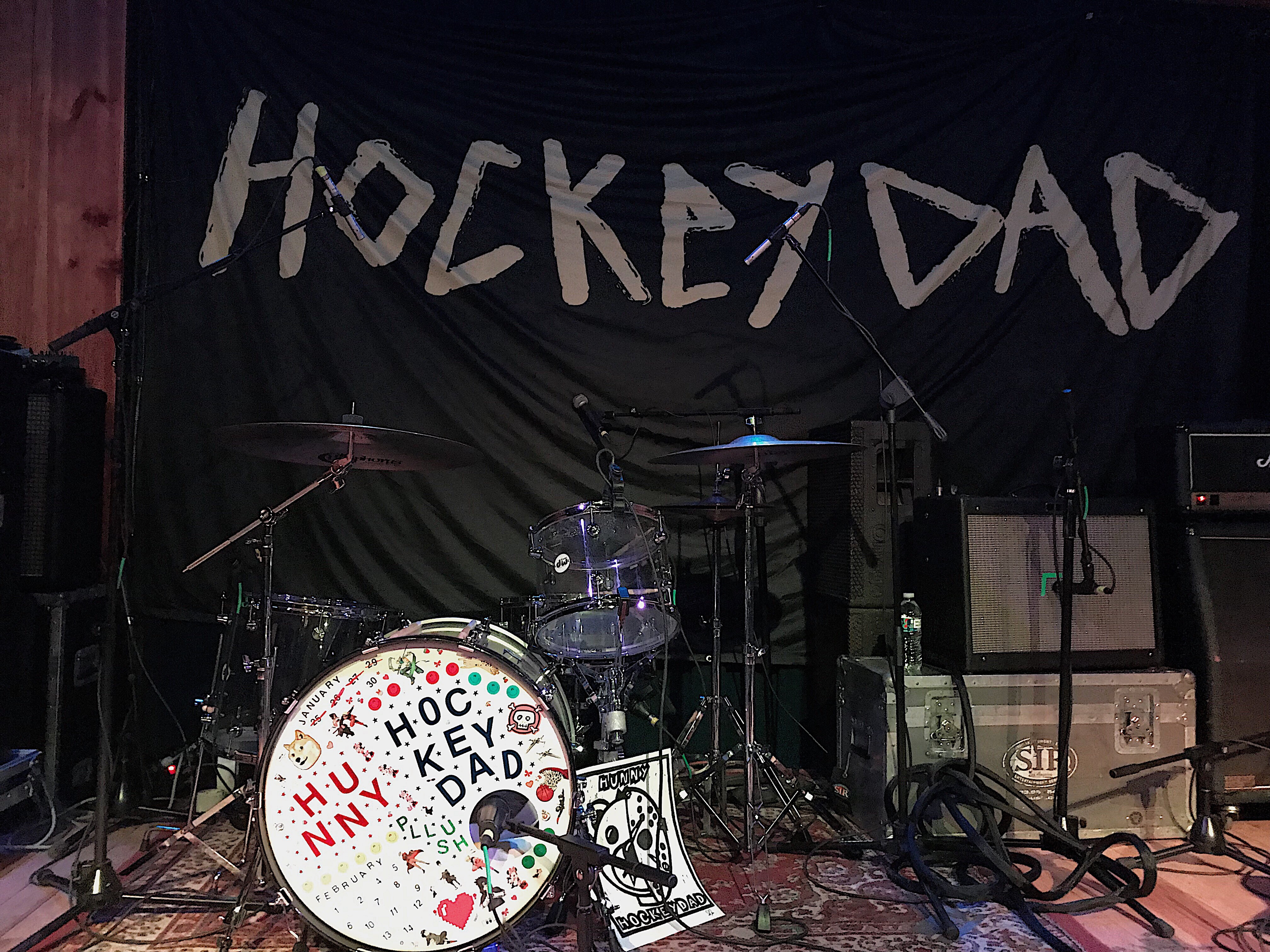A banner with "Hockey Dad" on it and a drum kit decorated with stickers spelling the band names.