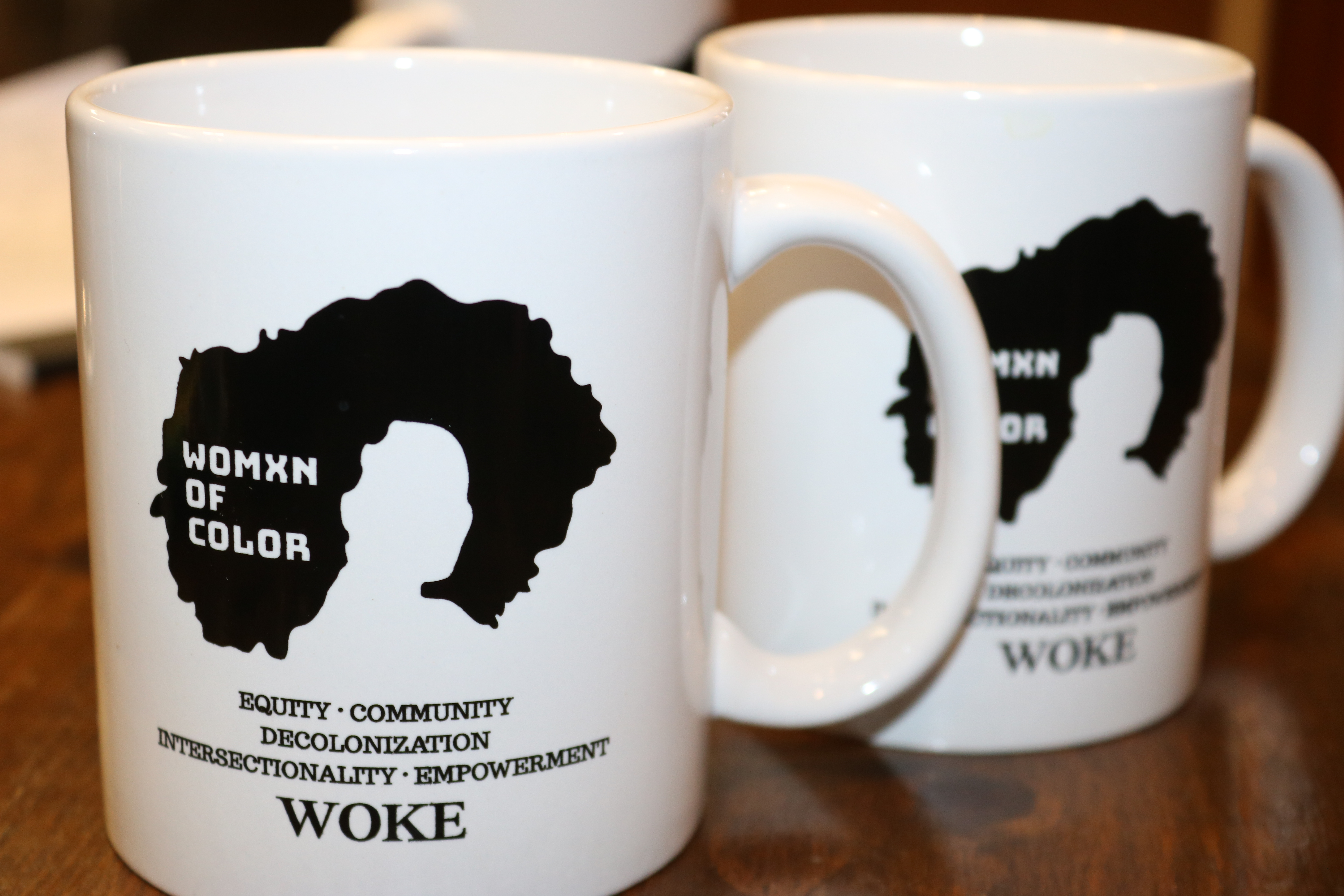 A white mug that shows a black outline of a woman’s afro. Beneath the outline are the words , Equity, Community, Decolonization, intersectionality, Empowerment and woke in all Caps.