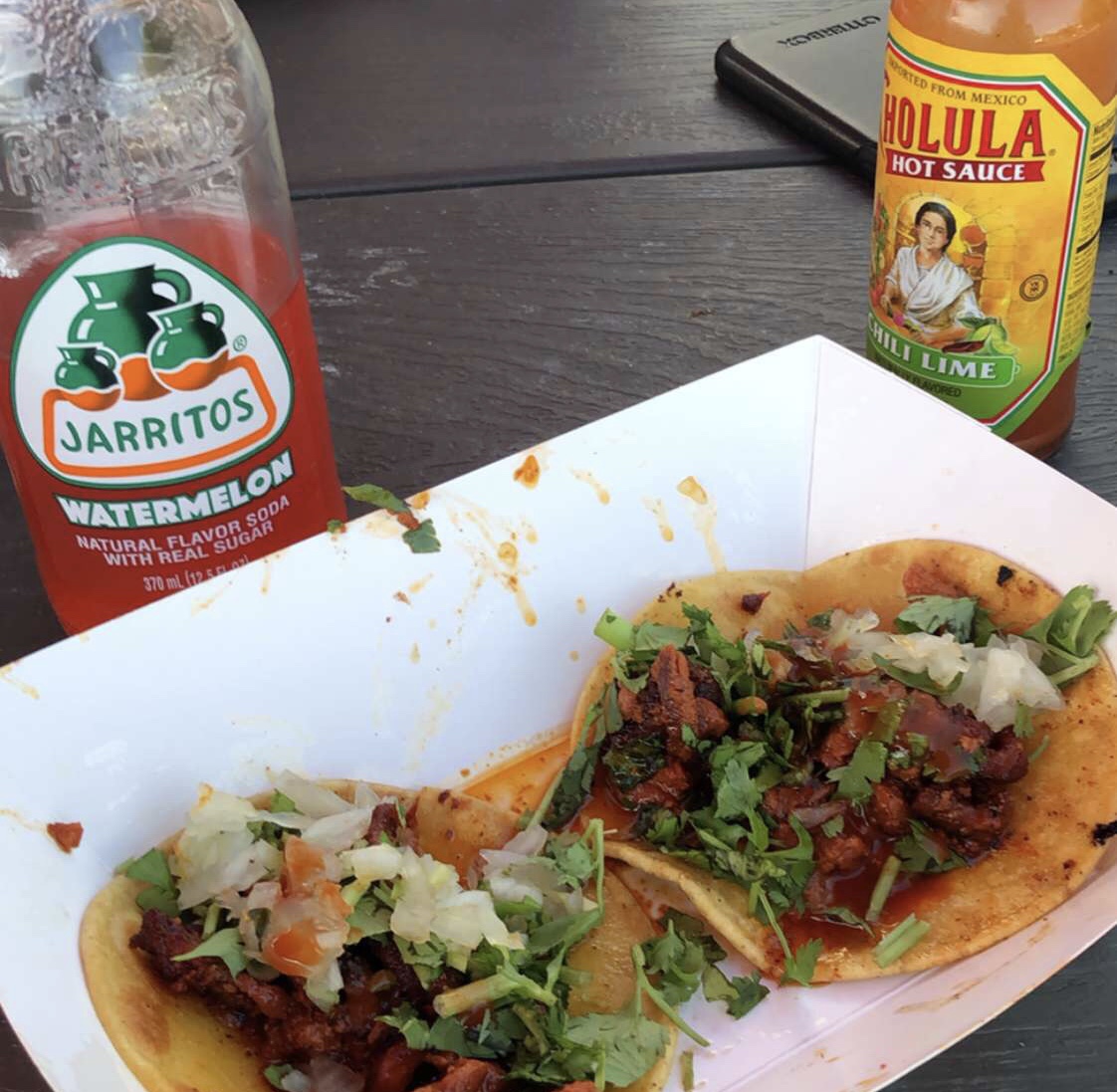 A pair of street tacos with a Jarritos and Cholula Hot Sauce bottle in the background.