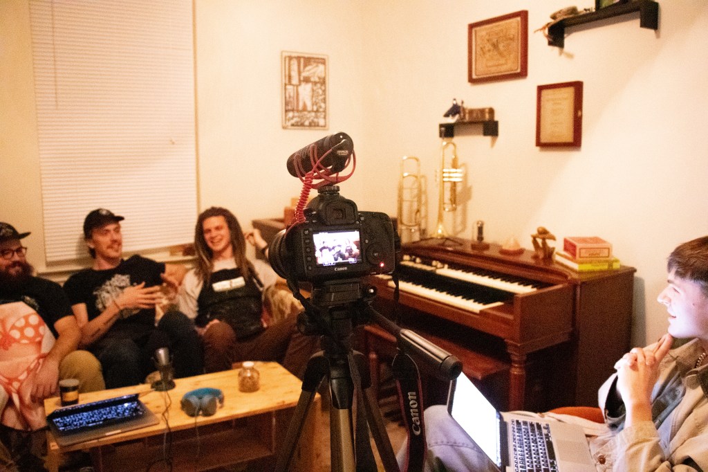 The three members of Kairos (Jake, Jacob, and Brent) sitting on a couch among music instruments and in front of the camera with interviewer, Conner Yarbrough, sitting behind the camera