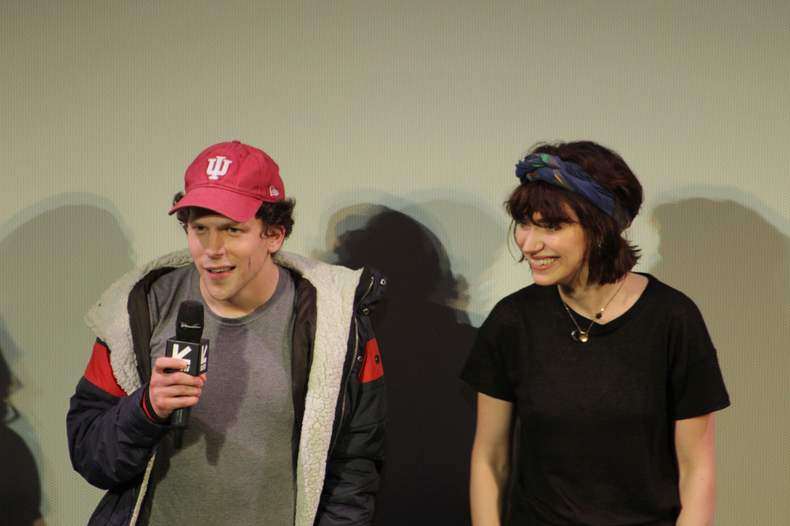 Jesse Eisenburg in a red cap and parka holding a microphone.