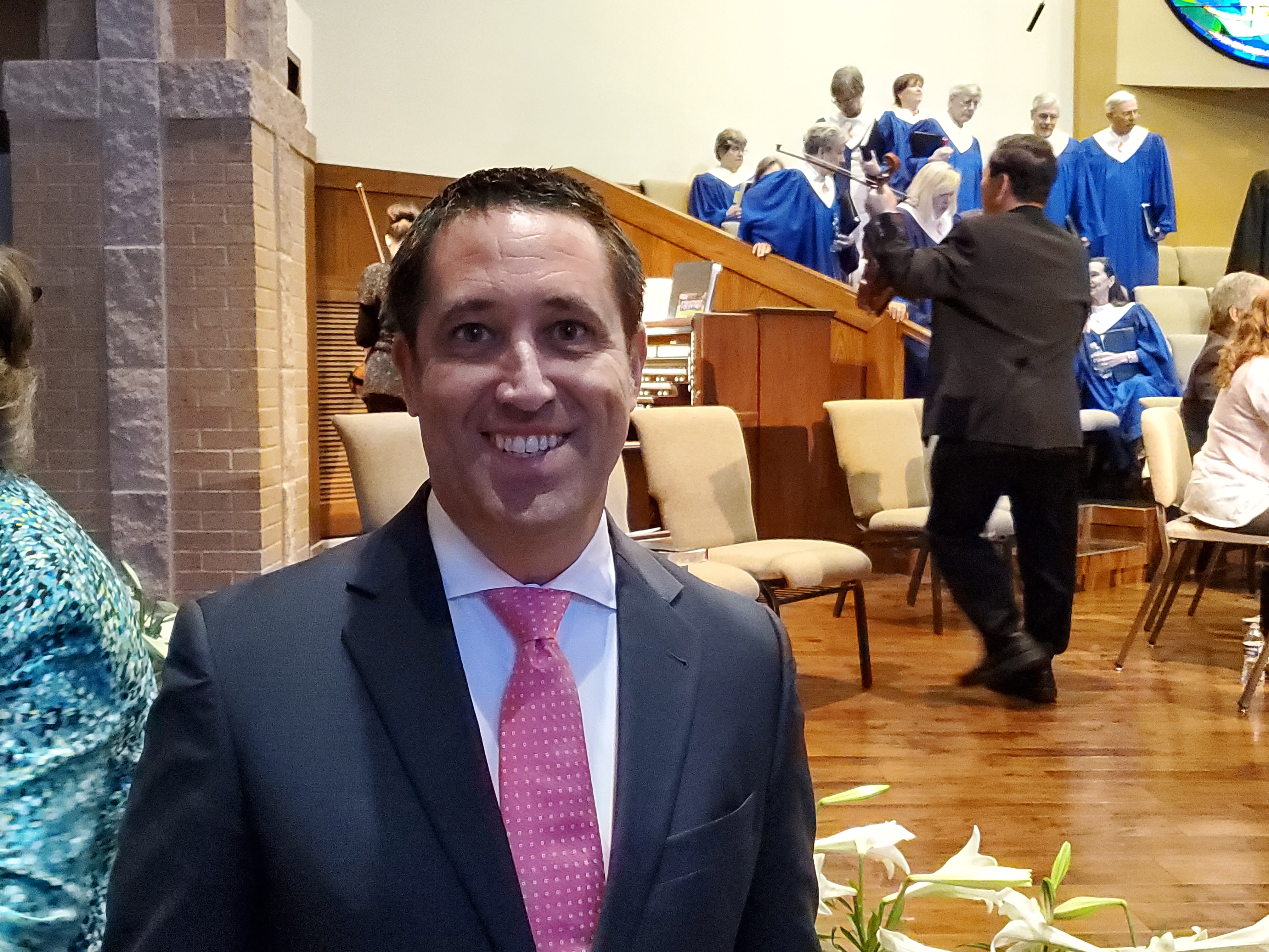 Glenn Hegar stands in front of the church altar. Several Choir members in robes can be seen in the background as well as other church attendees.
