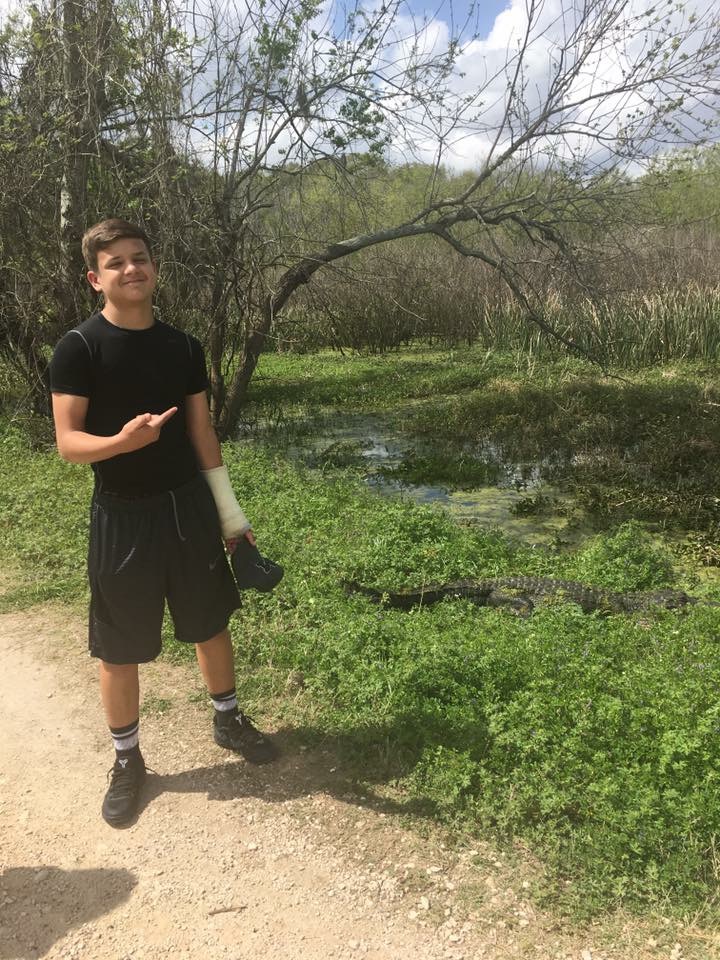 A teenage boy with an arm cast on pointing to the side where there is an alligator a few feet away in front of a swampy area off of a dirt walking trail