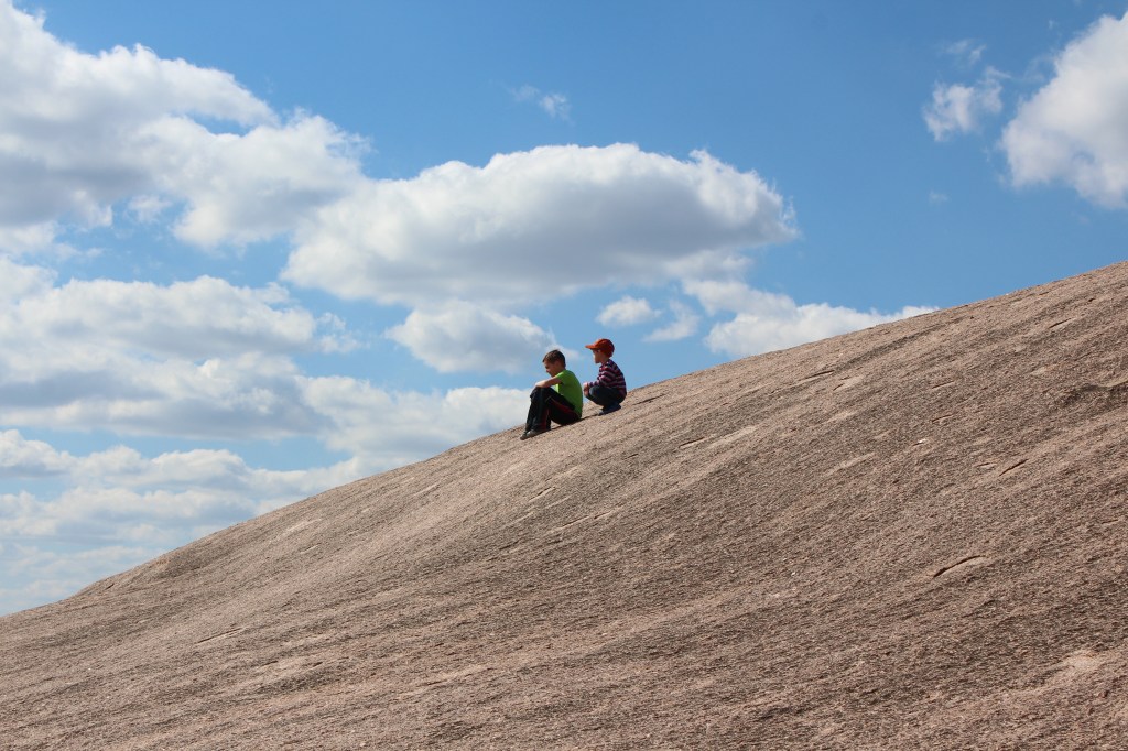 Two young boys sitting on the rock with the blue sky and clouds behind them.