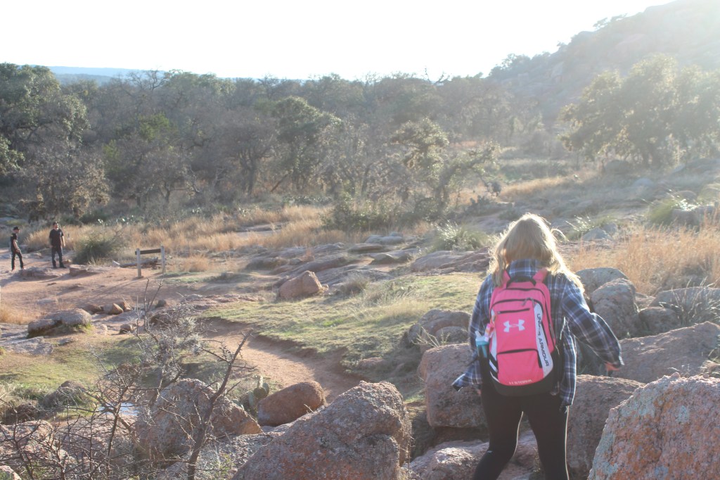 a girl wearing a flannel and a backpack with blonde hair walking amongst rocks as the sun lowers