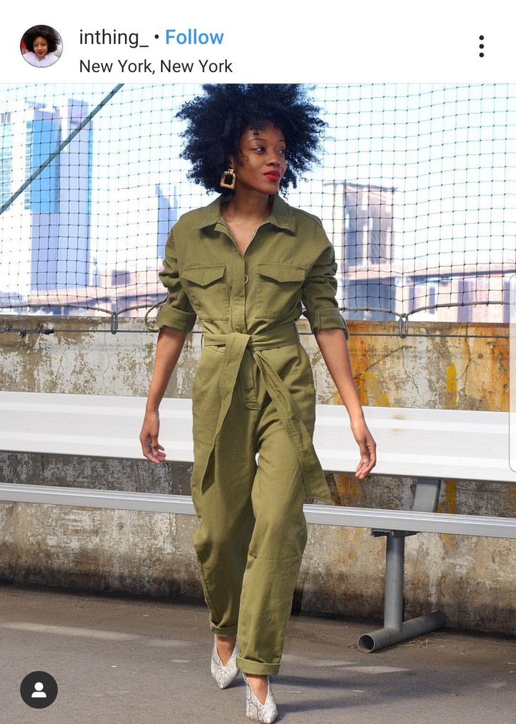A woman wearing a green boiler suit with a fence background, with buildings in the distance.
