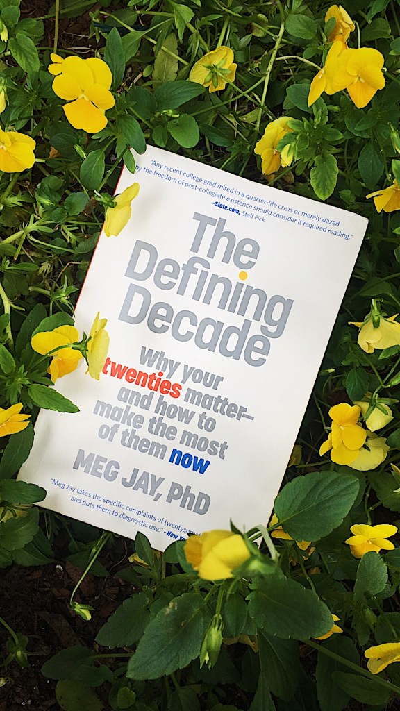 A copy of “The Defining Decade” by Dr. Meg Jay laid on top of yellow flowers.