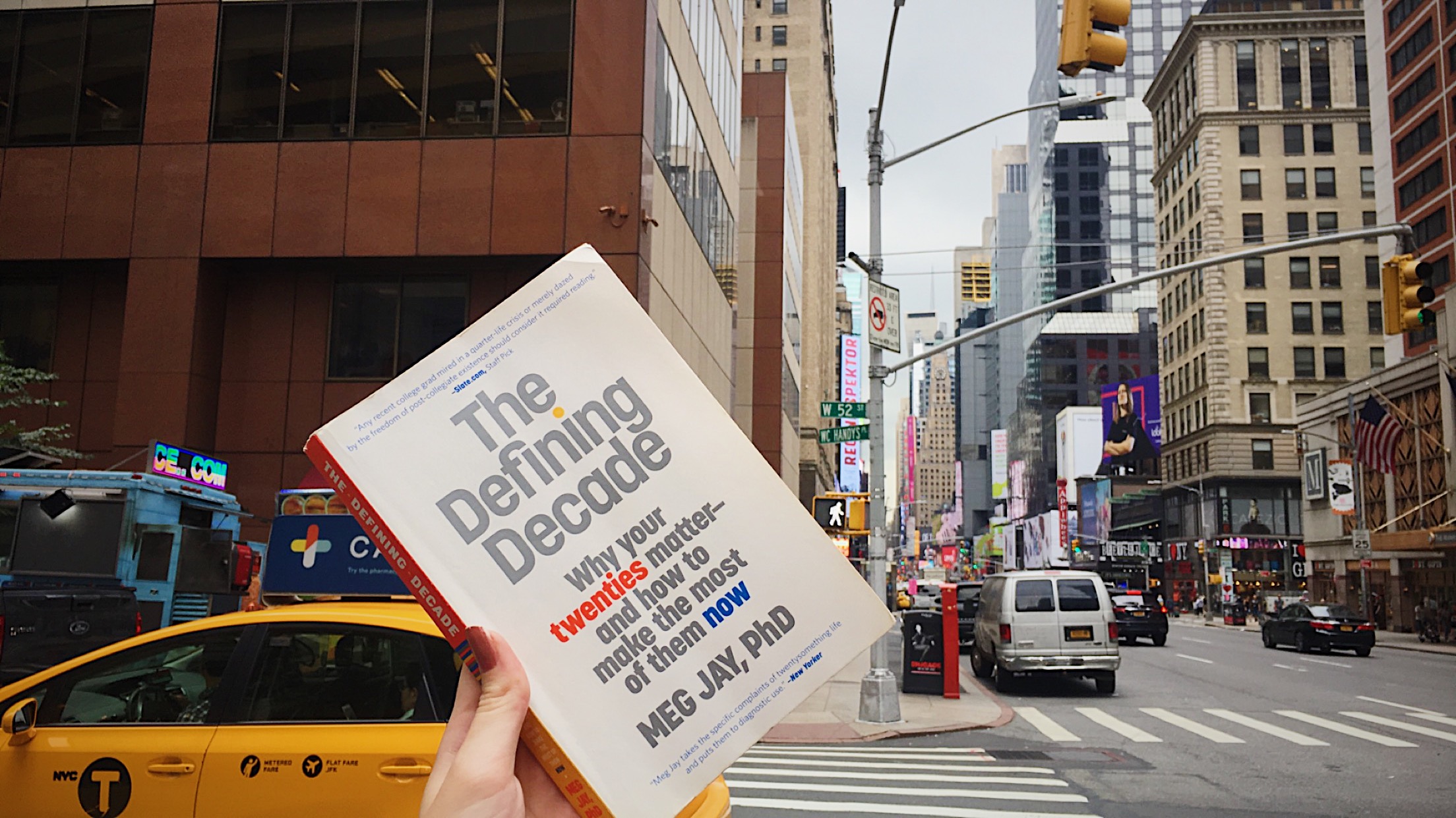 A copy of “The Defining Decade” by Dr. Meg Jay held up from a New York street against Times Square buildings.
