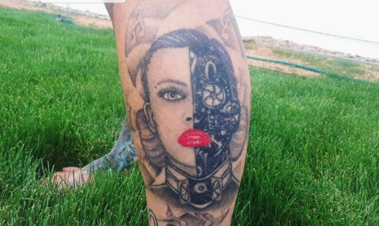 A tattoo of a woman’s head on the outer calf area of a woman’s leg. Half of the head is gears and metalwork, and the other half is normal, and the woman is wearing bright red lipstick and has purple hair pulled into two buns on either side of her head.