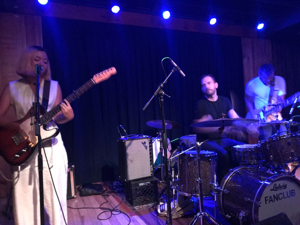 Three people on stage. Left to right; one girl playing guitar and singing, one guy playing drums, and the last guy playing bass.