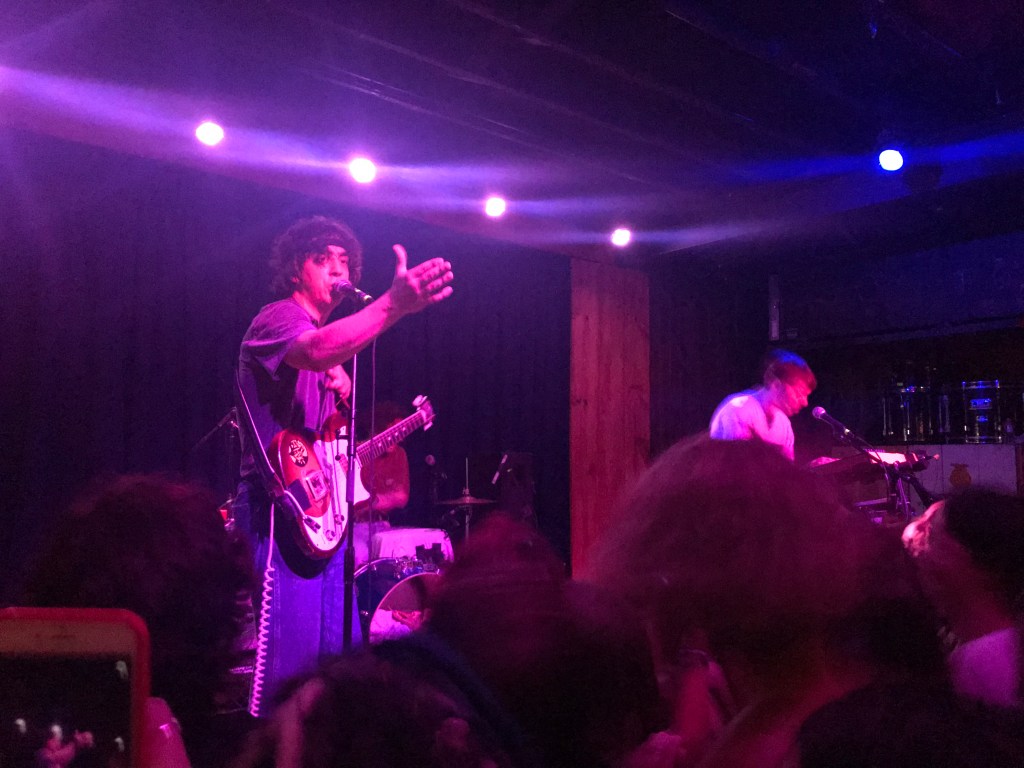 Two guys on stage, one with curly dark hair playing guitar and singing and reaching out to the audience, the other with straight light hair singing and playing the synth.