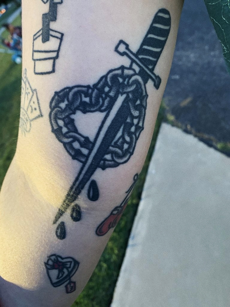 A tattoo on a forearm of a dagger going through a heart made of chains, with three drops coming off of the end of the dagger.