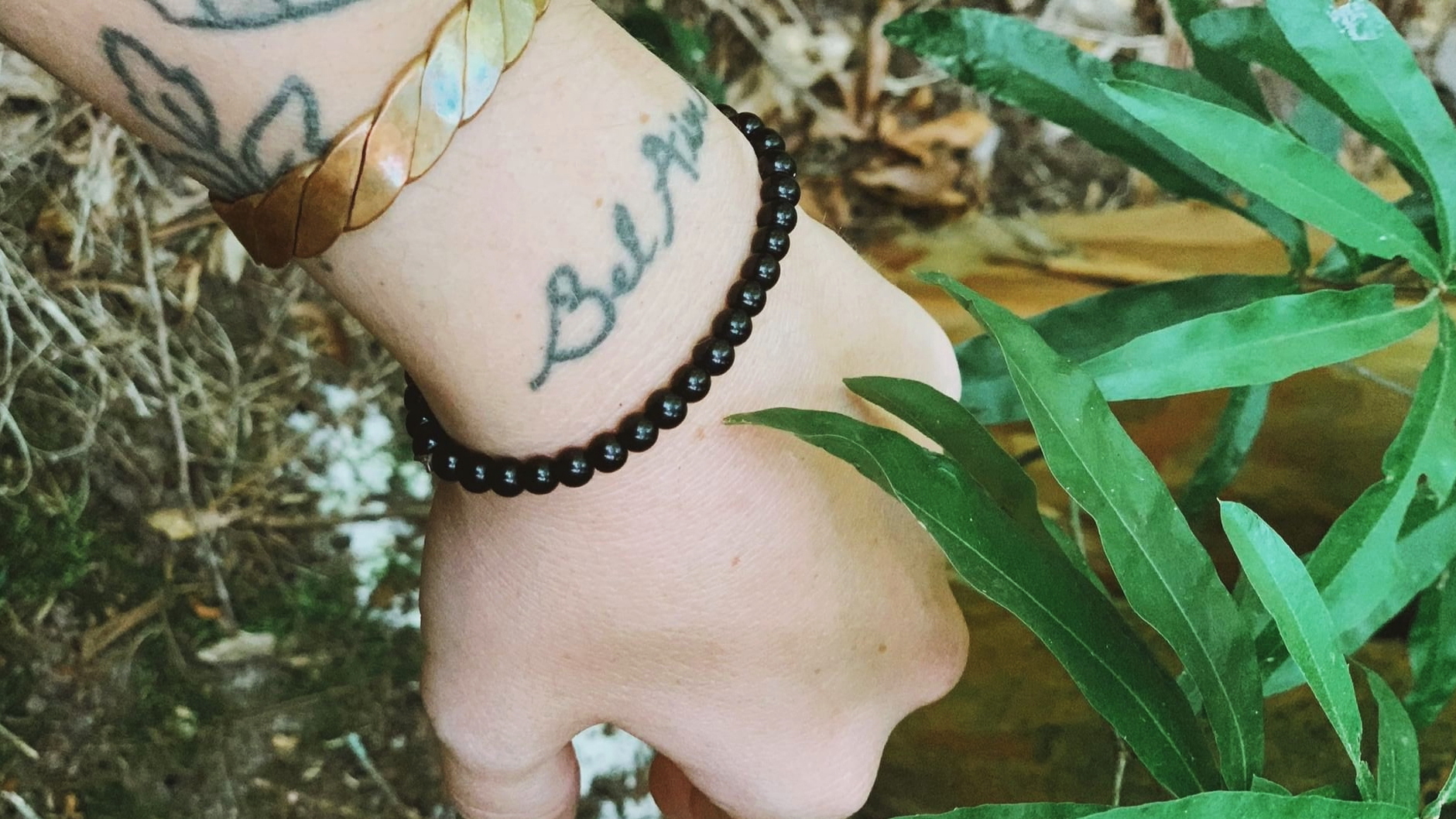 A tattoo with the words “Bel Air” written in cursive on the top of a forearm. The arm has a black beaded bracelet and a gold bracelet and there are grass and leaves in the background.