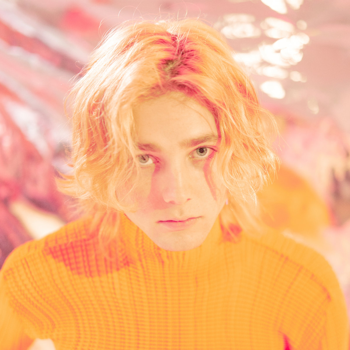 he album cover is a headshot of Instupendo. He’s wearing an orange fitted turtleneck and has blonde hair. Behind him is a pink and silver backdrop.