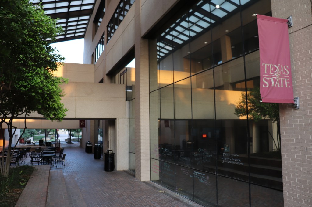 The image gives an idea of where the writing center is located. It shows the Writing Center windows to the left and includes the outdoor tables straight ahead. On the far left a Texas State University flag is hung from the building.