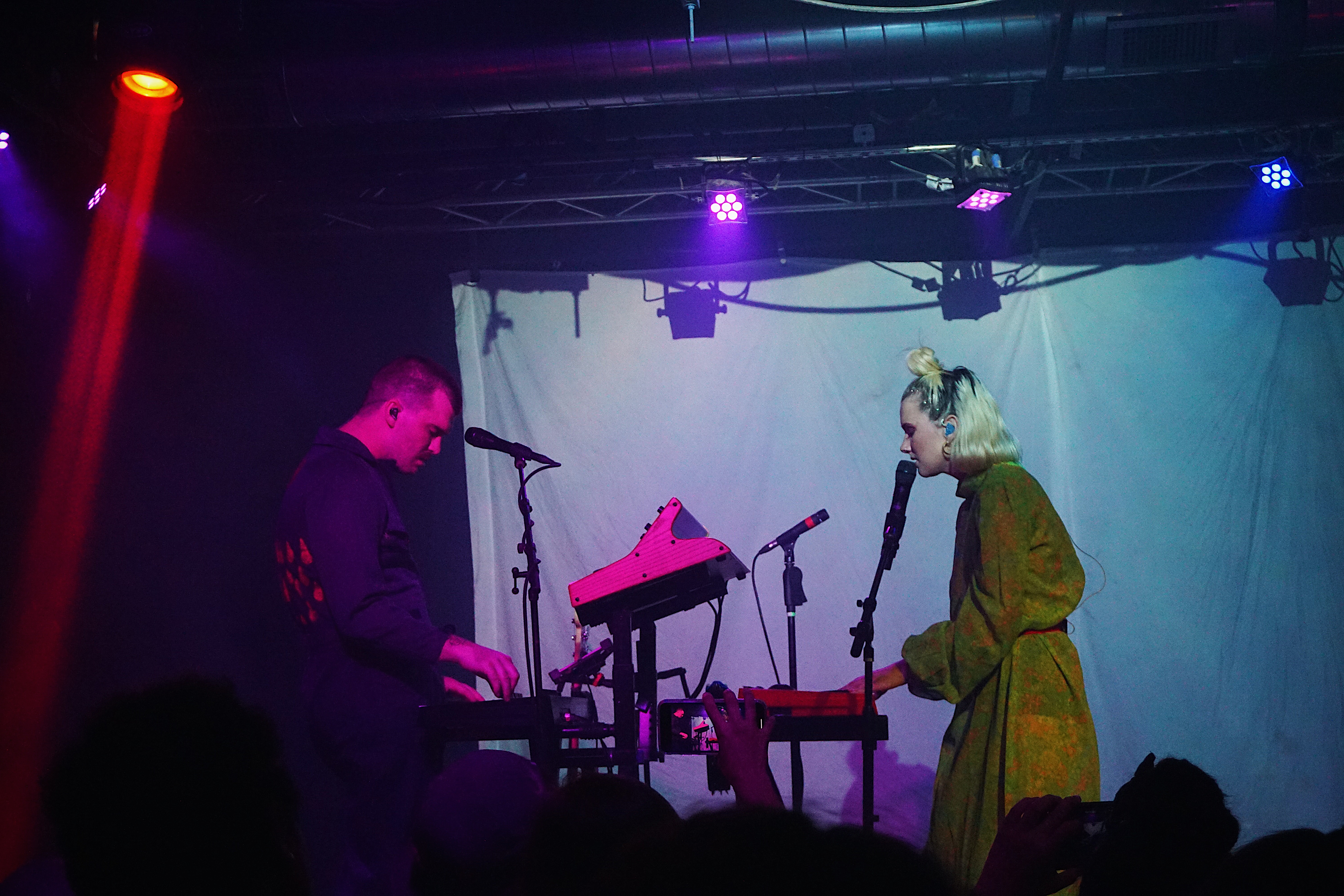 A man and a woman face each other on a concert stage while both playing a keyboard. The picture is dark with the stage lit up by artificial light.