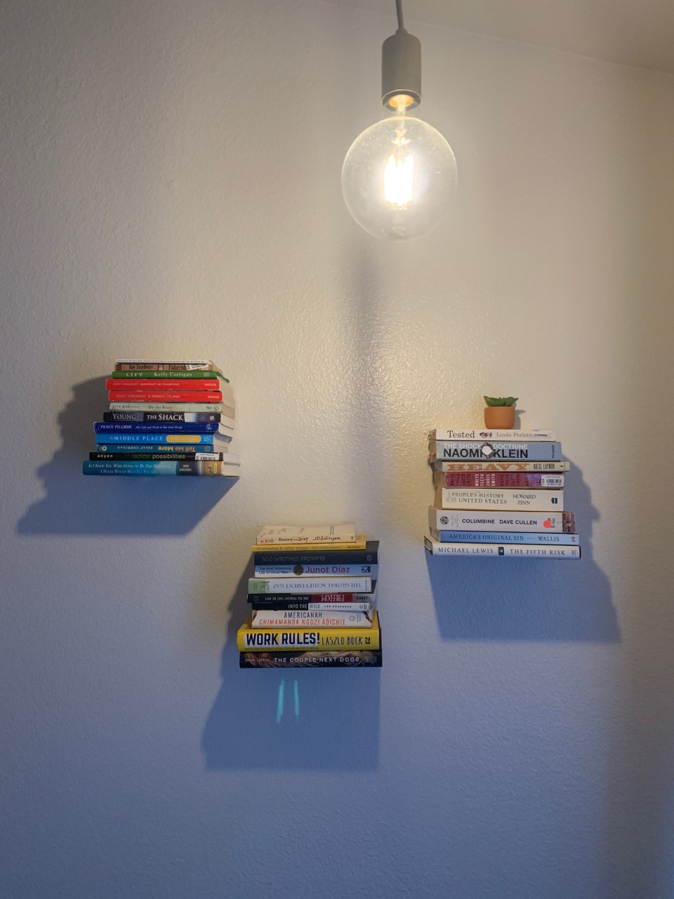 Three stacks of books on three floating shelves on a white wall with a large light bulb