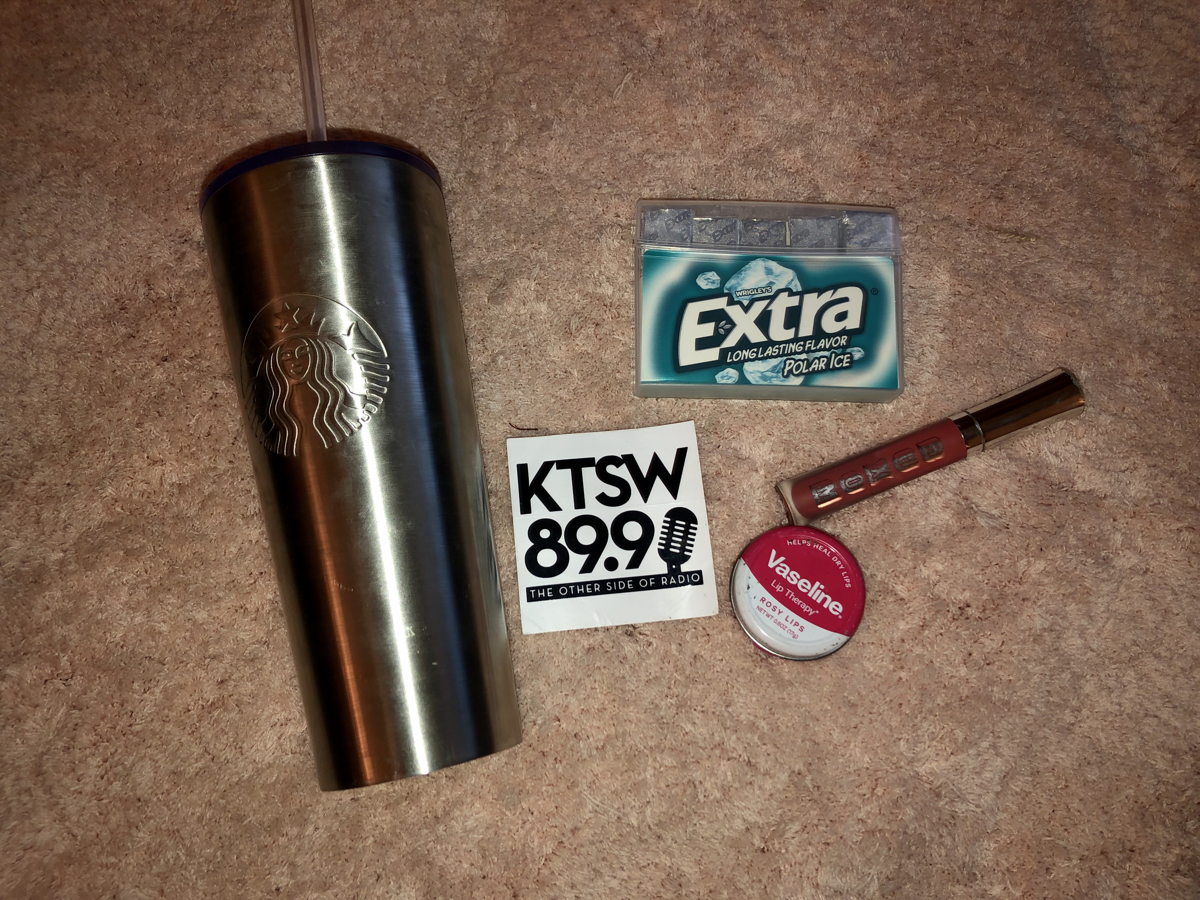 A silver Starbucks cup next to a white KTSW sticker, blue gum package, red lipgloss and pink chapstick
