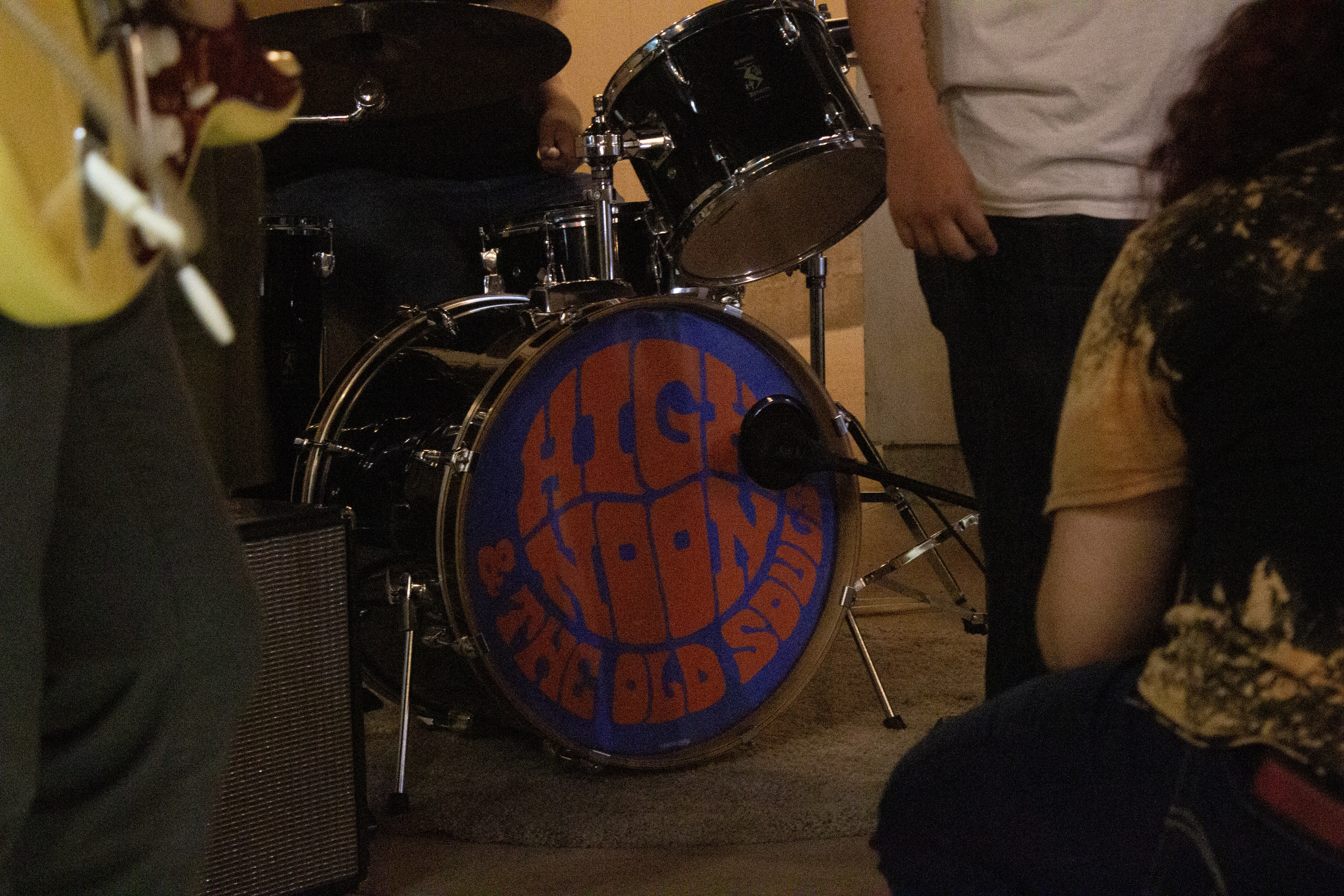 the photo is centered on the bass drum of the drum kist, featuring a logo of the band’s name, High Noon and the Old Souls. Band members are gathered around the kit, setting up their respective instruments