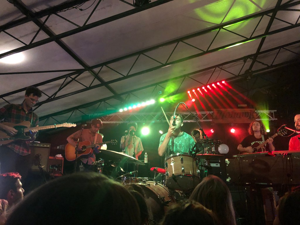 The picture shows all members playing their instruments (3 guitarists, one bassist, and two keyboardists) while the drummer sings into a microphone with a single drumstick in his left hand.
