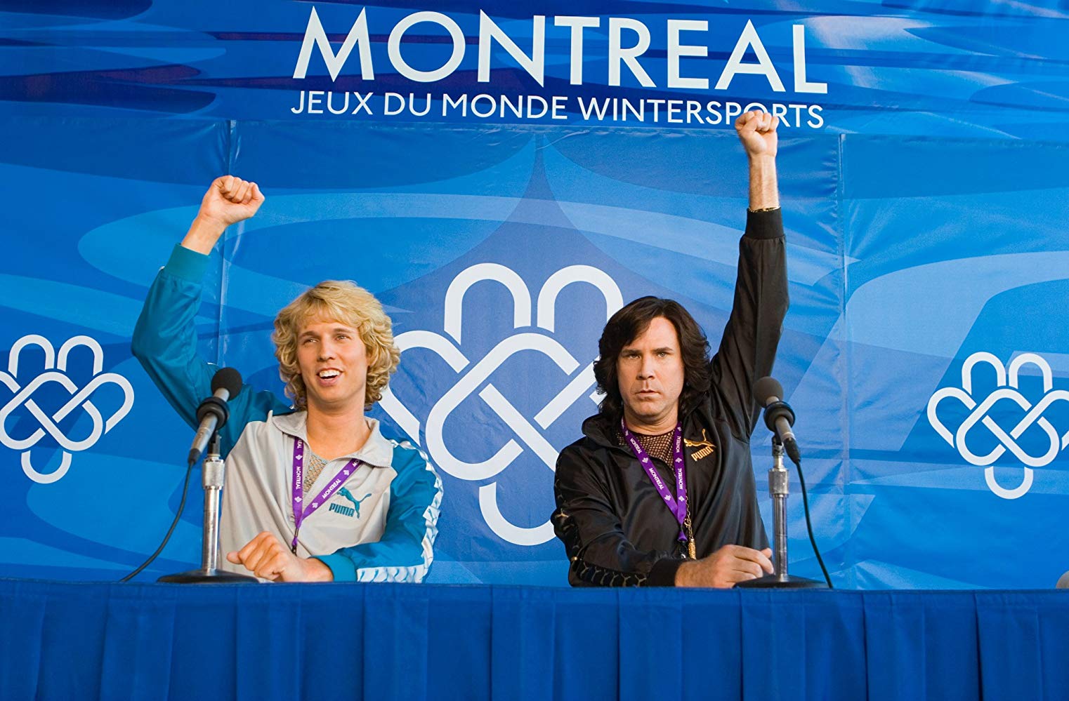 Two men on a panel at a press conference after a figure skating competition, both holding their fists in the air with the title “Montreal Jeux De Monde Wintersports” displayed behind them.