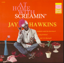 Singer Screamin’ Jay Hawkins sits Indian style against an orange background. He is surrounded by vases and flowers and wears a white and purple hat and sunglasses. The words “At Home with Screamin’ Jay Hawkins” reads in large font behind him.