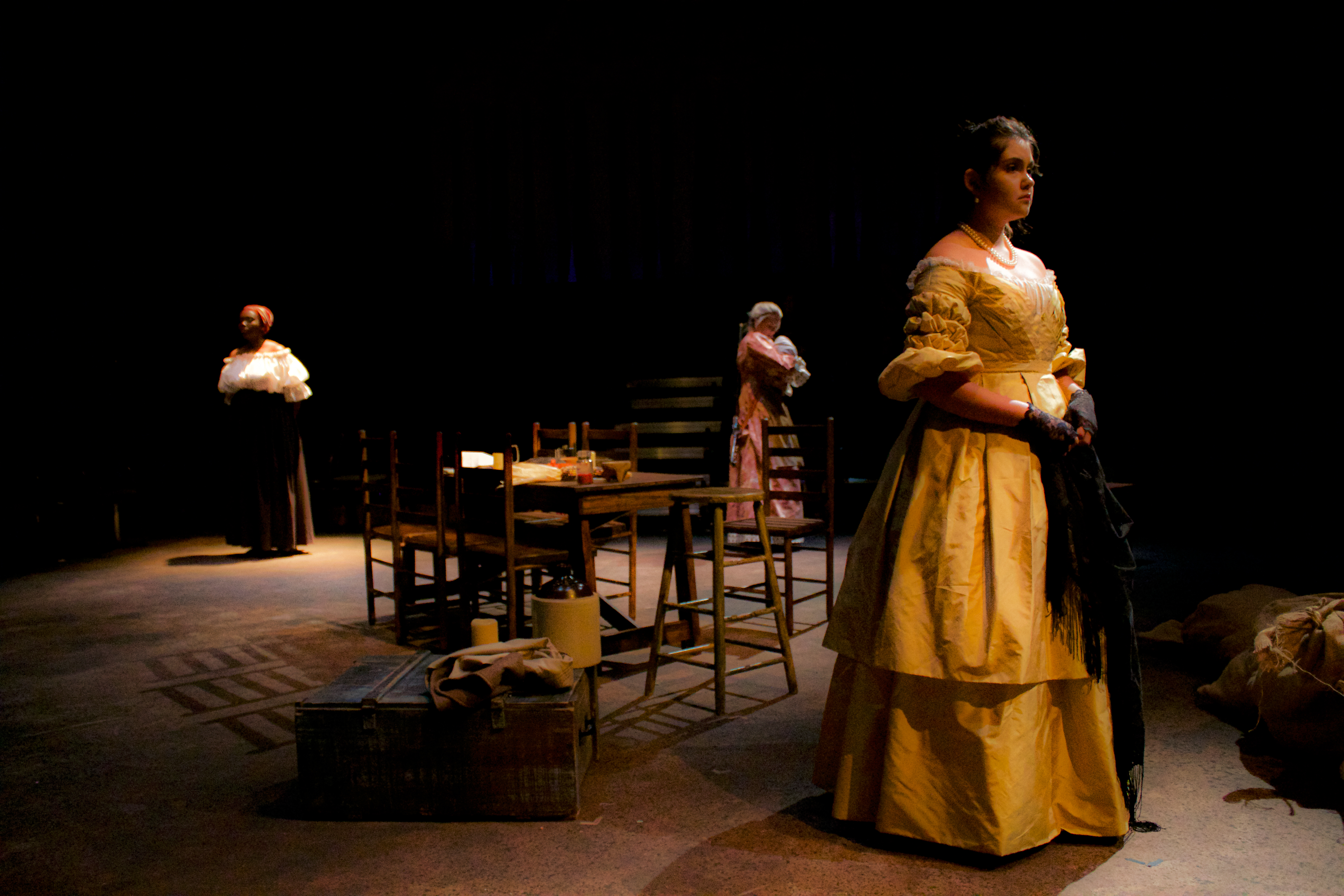 Three women standing in different corners of the stage with a table between them.