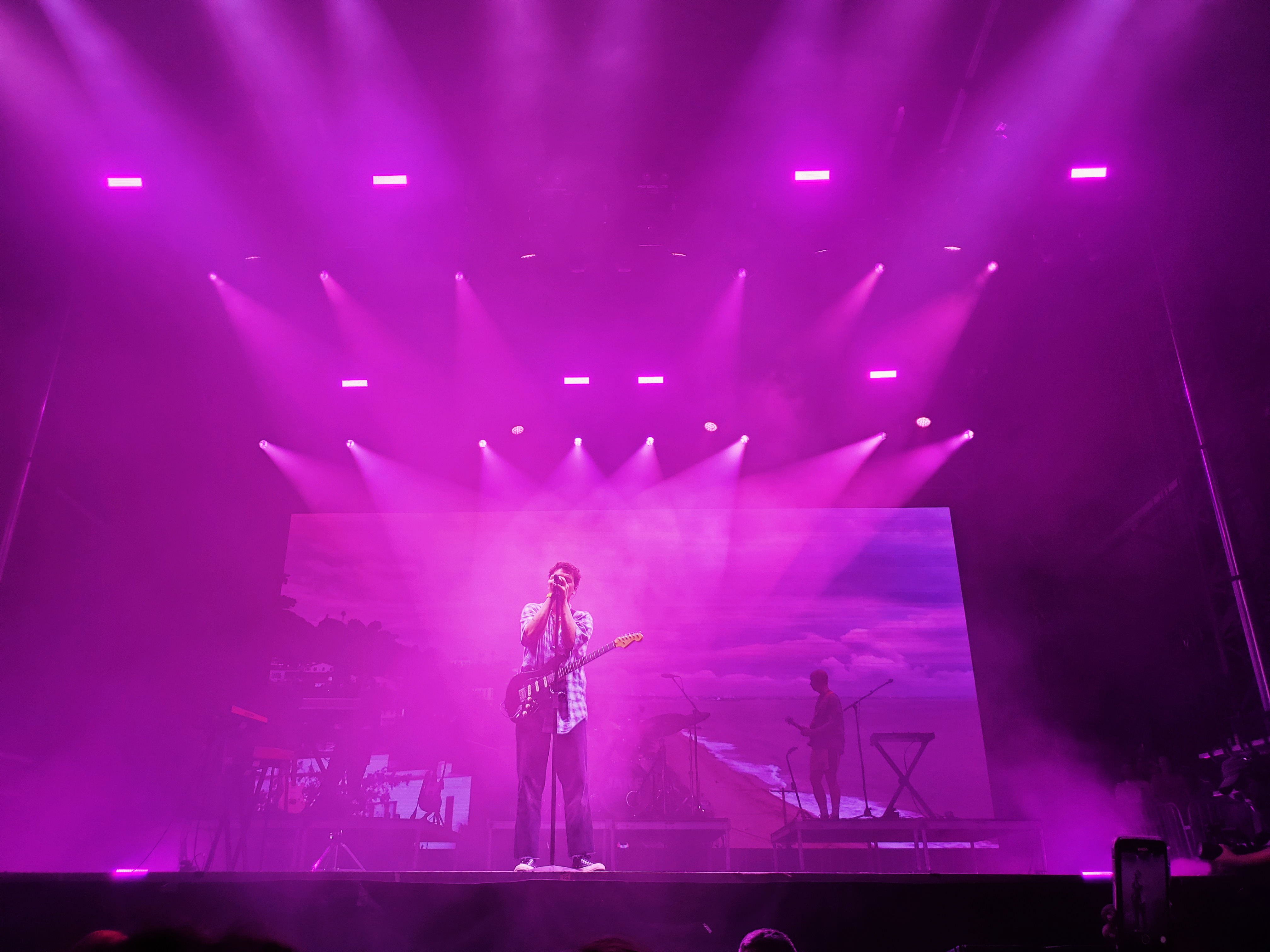 LANY performing at Austin City Limits. Paul Klein is in the center at the microphone. The background is a beach with a cloudy sky and the spotlights make the stage pink.