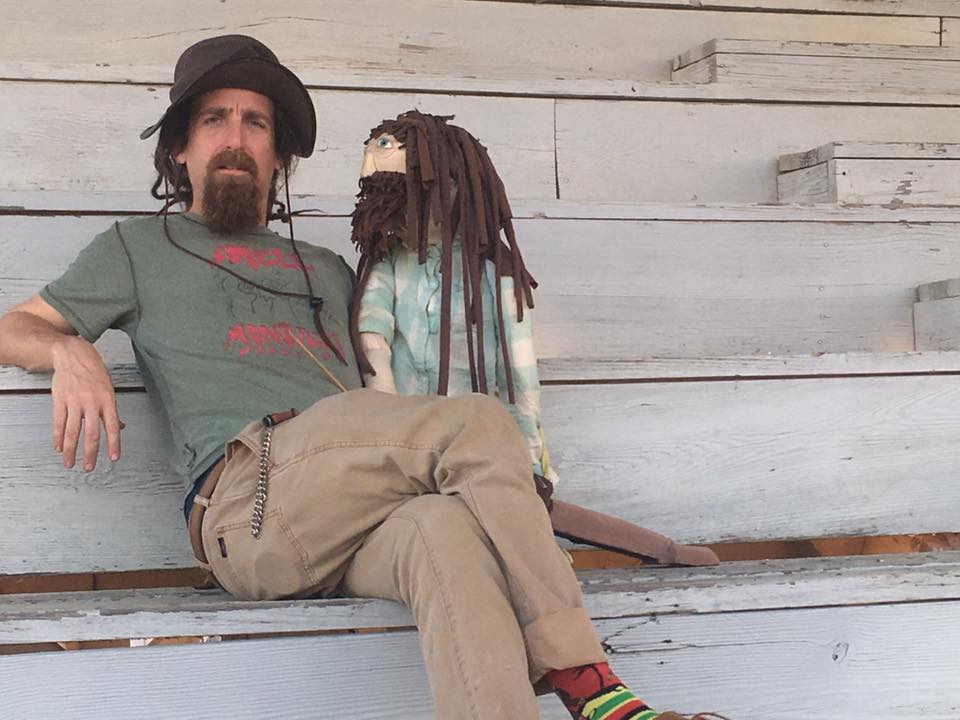 A man wearing a hat, with a brown beard and dreadlocks, sits on a step wearing a shirt that says “Smell Monsters Puppetry” and holding a puppet version of himself.