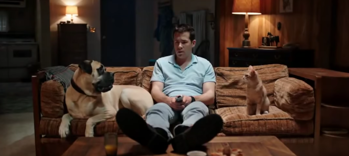 Jerry wears a blue polo shirt while sitting on his brown couch in between his dog and cat.