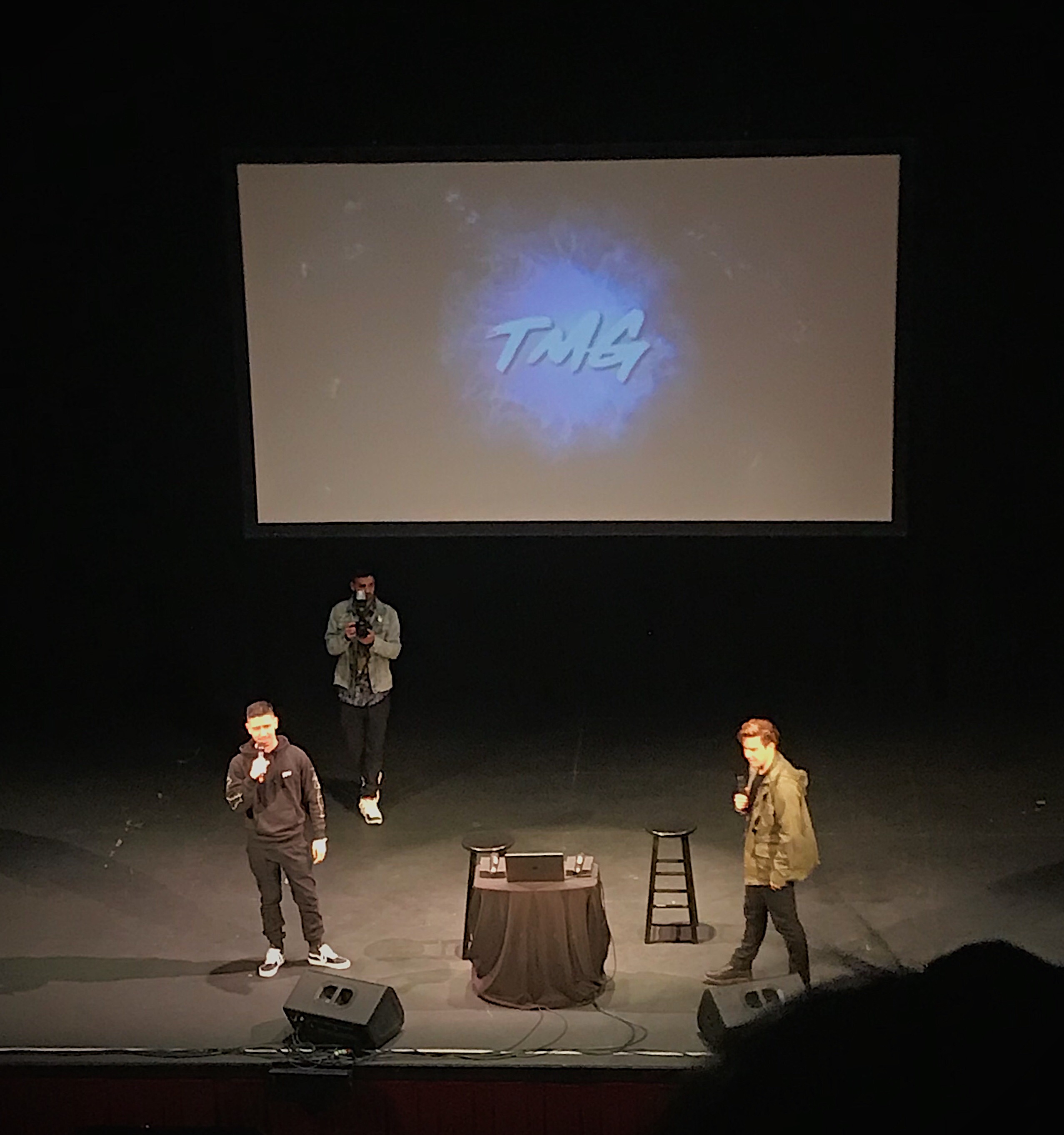 Noel Miller stands left of Cody Ko on a theater stage. They are both spotlighted by overhead lights. A projector screen above them reads “TMG” on top of a tye-dye blue background.