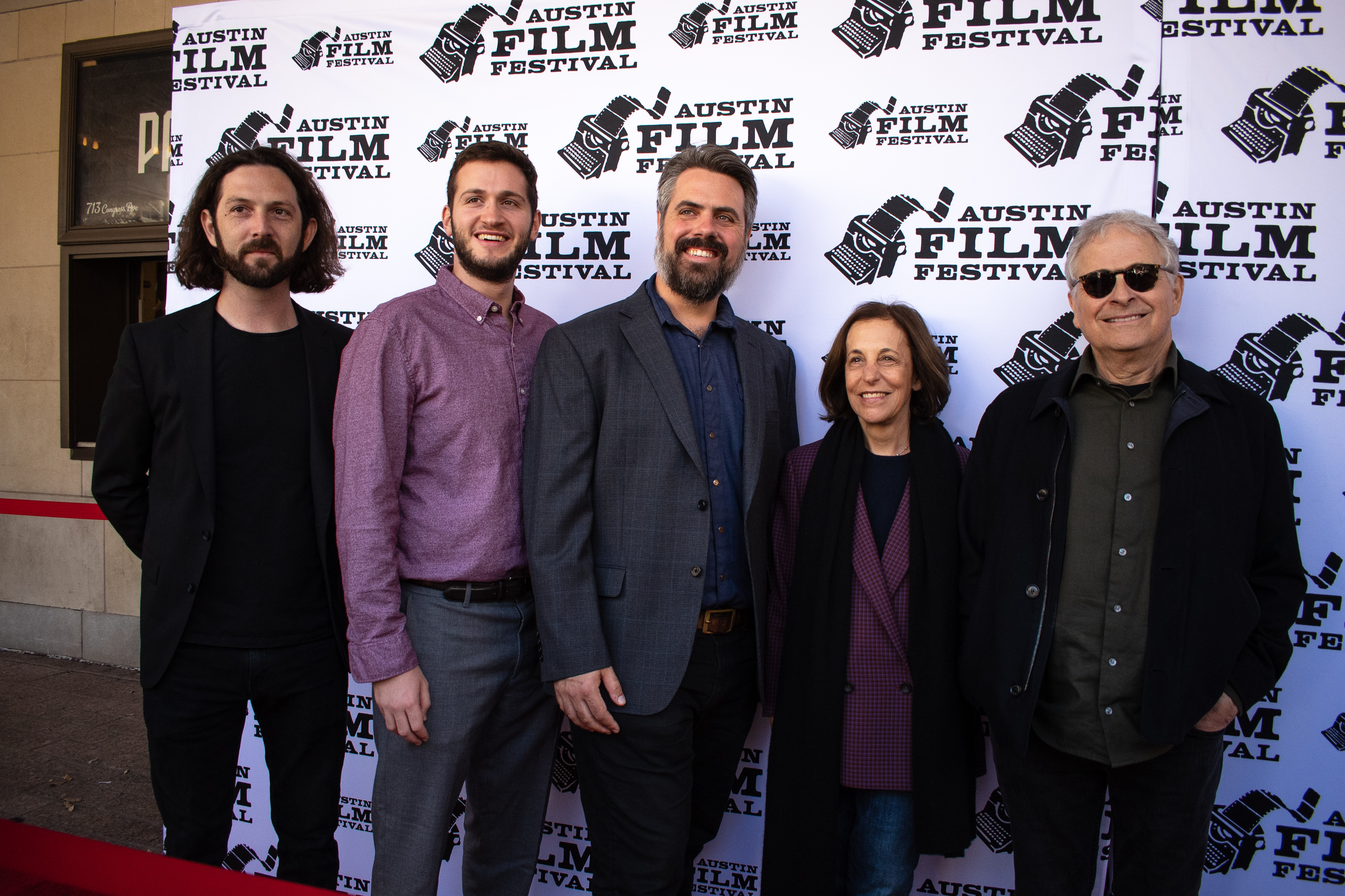 3 men stand next to eachother on the left and a woman stands in between the three men and one man on her right. They are all posing for a picture on the red carpet.