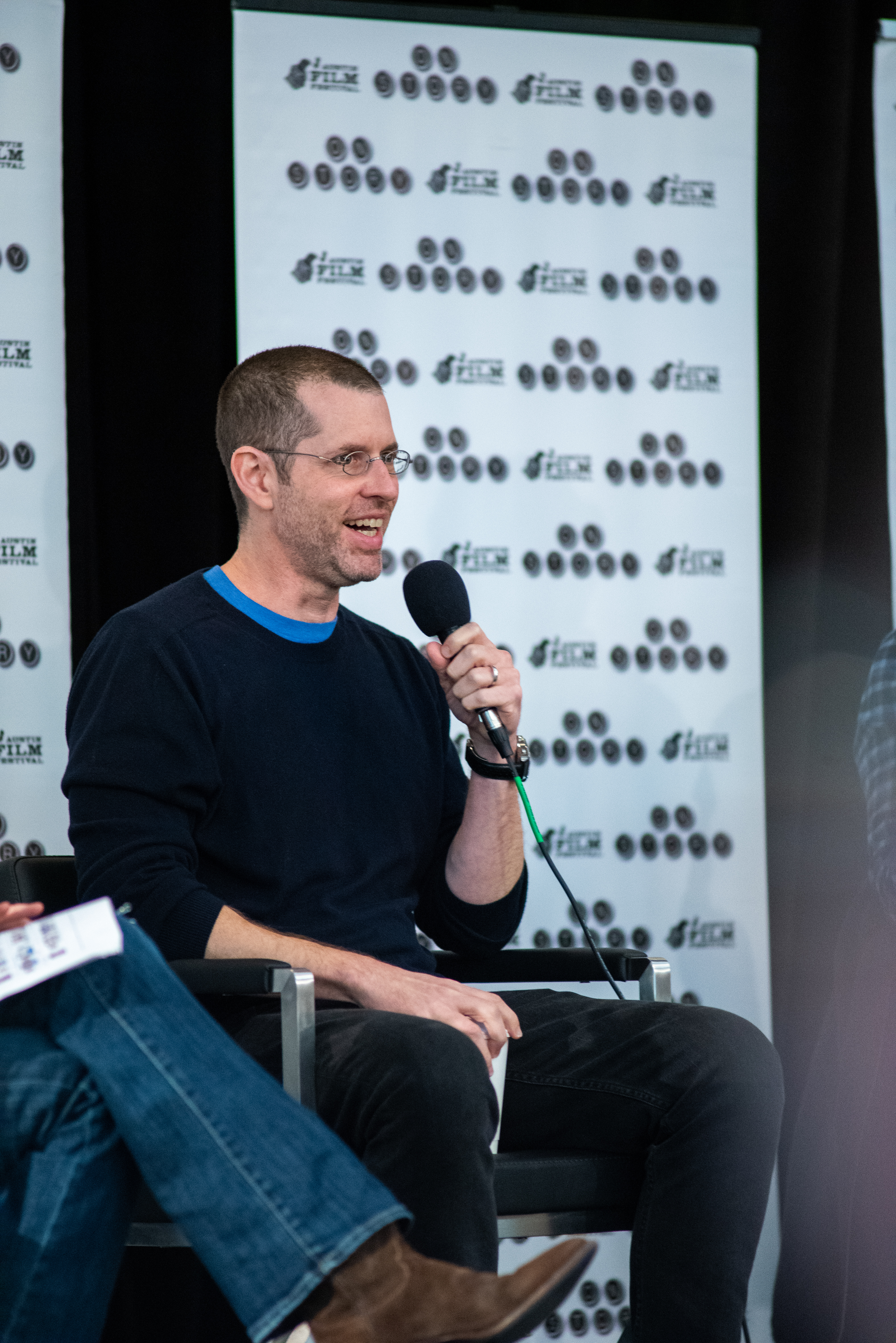 Man sitting in a chair on stage speaking into a microphone.