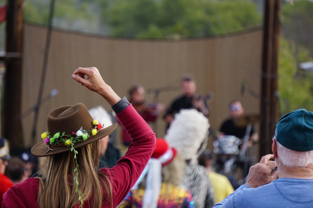 A woman in a brown hat wrapped with flowers puts her right hand up while listening to the Jon Stickley Trio