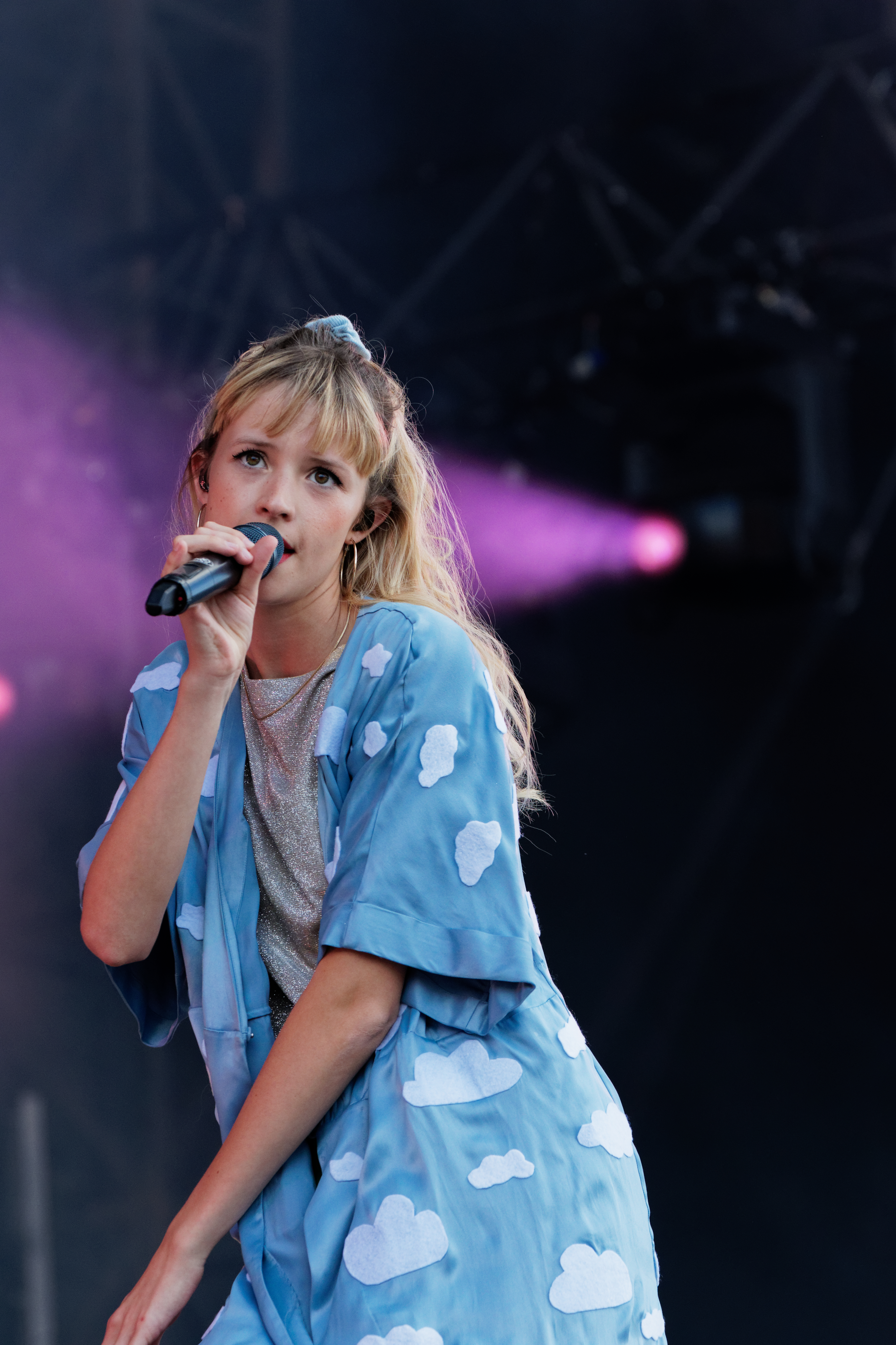 Artist Angèle wearing a blue, cloud patterned button down shirt performing on a stage holding a microphone up to her mouth while singing. She has long blonde hair pulled halfway up by a blue scrunchie with bangs.