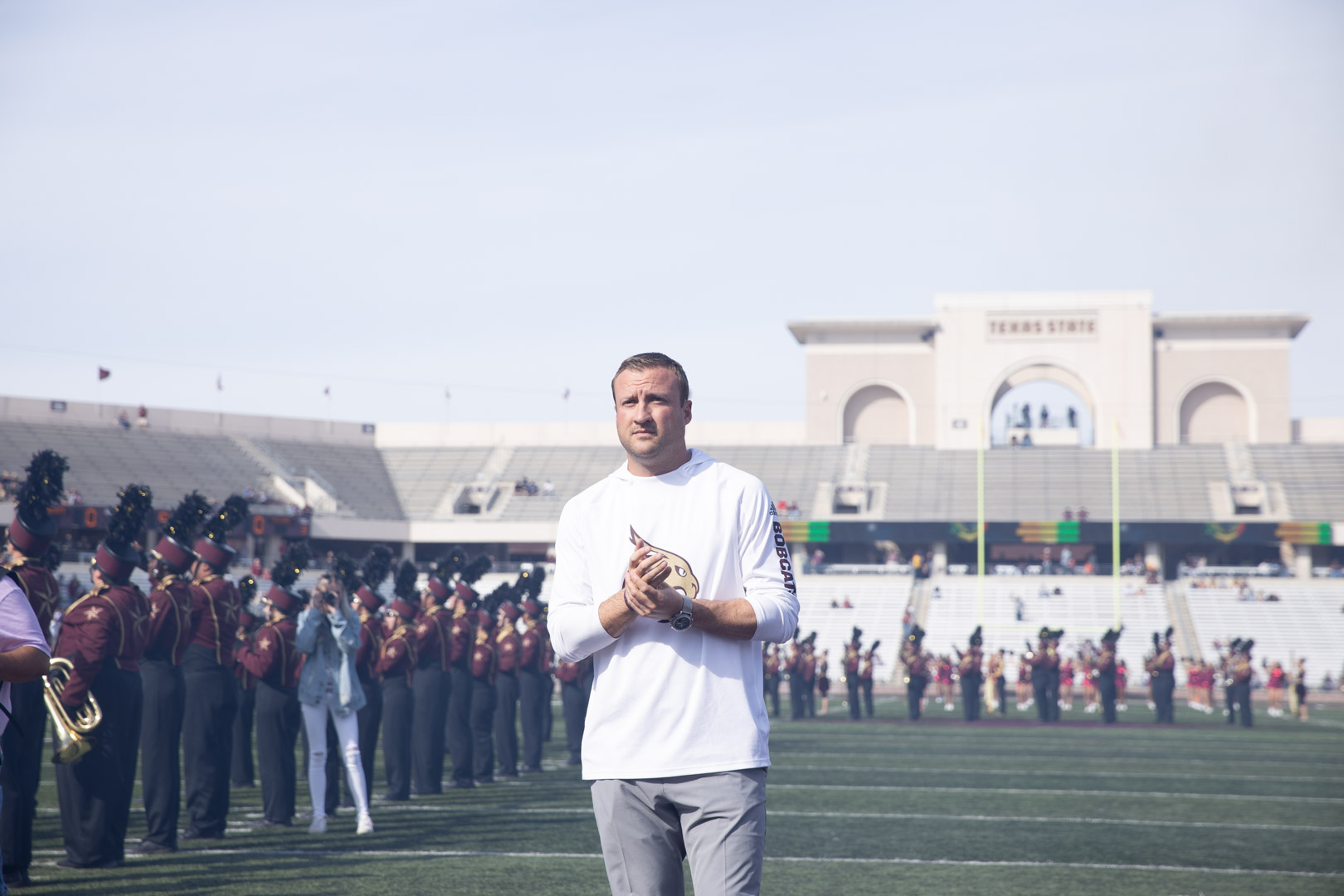 Jake Spavital stands on the field, looking towards the camera at the end zone complex with the Texas State Marching Band lines the field behind him.