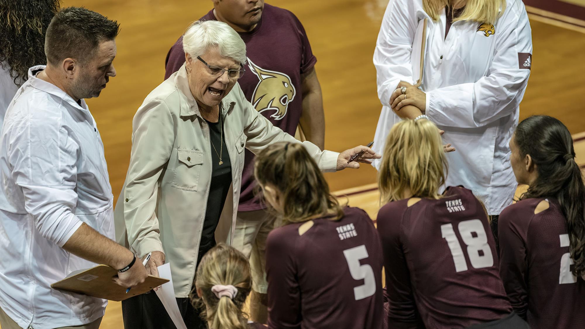 Karen Chisum and her assistant coaches stand in front of four Bobcat volleyball players in maroon jerseys with white lettering and numbers on the sideline in a match at Strahan Arena.