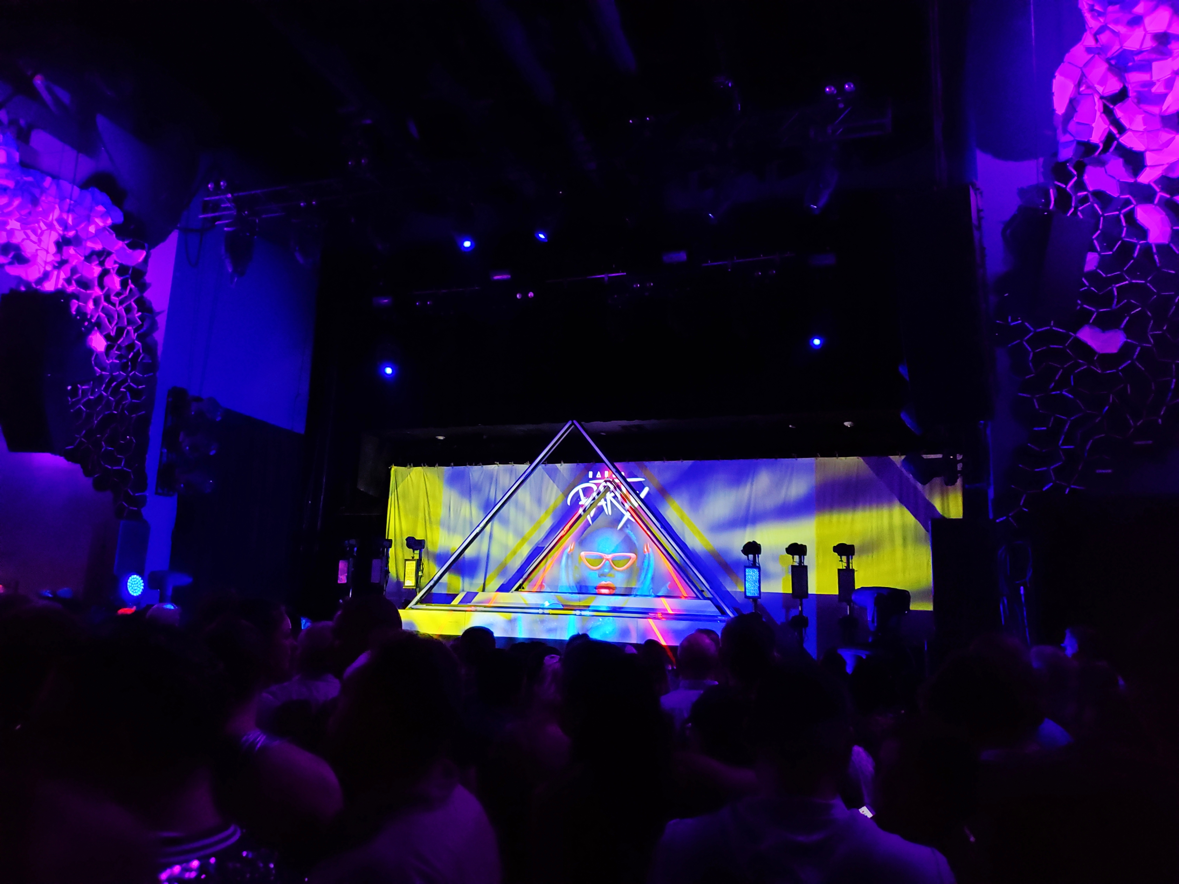 A picture of Emo’s stage. The lighting is purple, and the backdrop on the stage in a picture of Todrick Hall with red lips and sunglasses with the words “Haus Party” written above his head. There are two triangular fixtures on the stage.