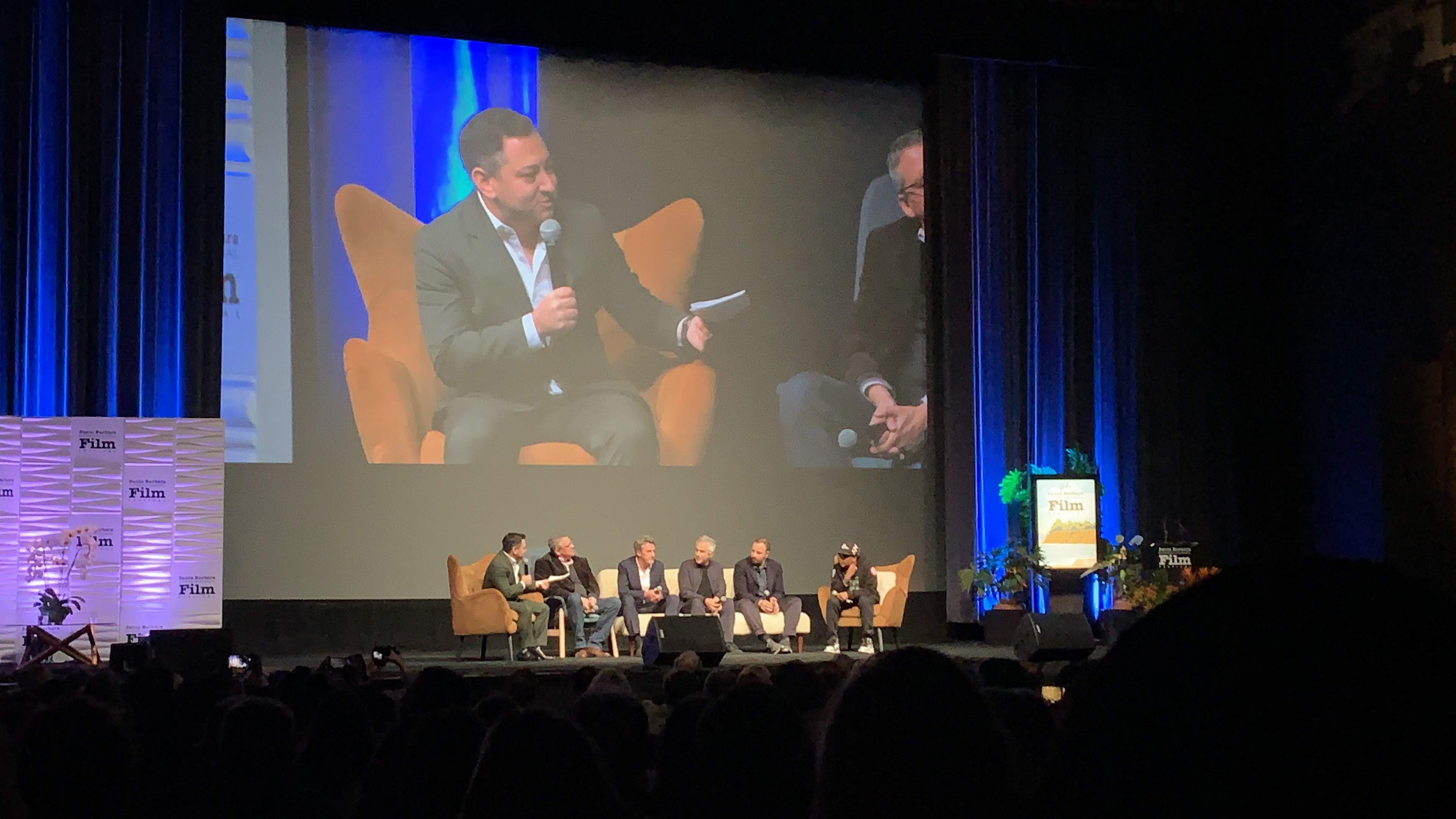 six men sitting in chairs on stage inside a theater