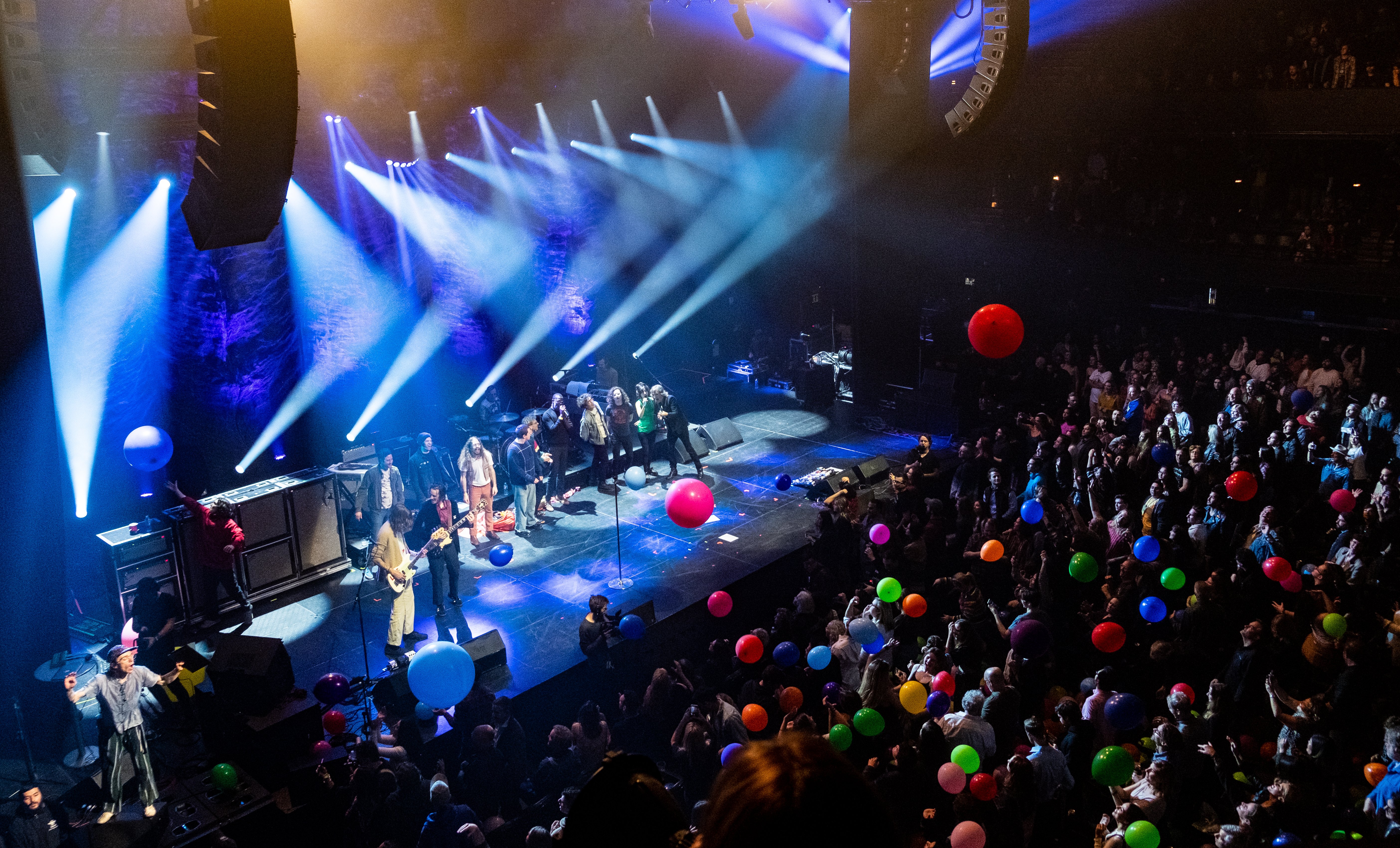 People on stage are singing and dancing. The crowd is playing with a dozen balloons, which are all different colors and sizes.
