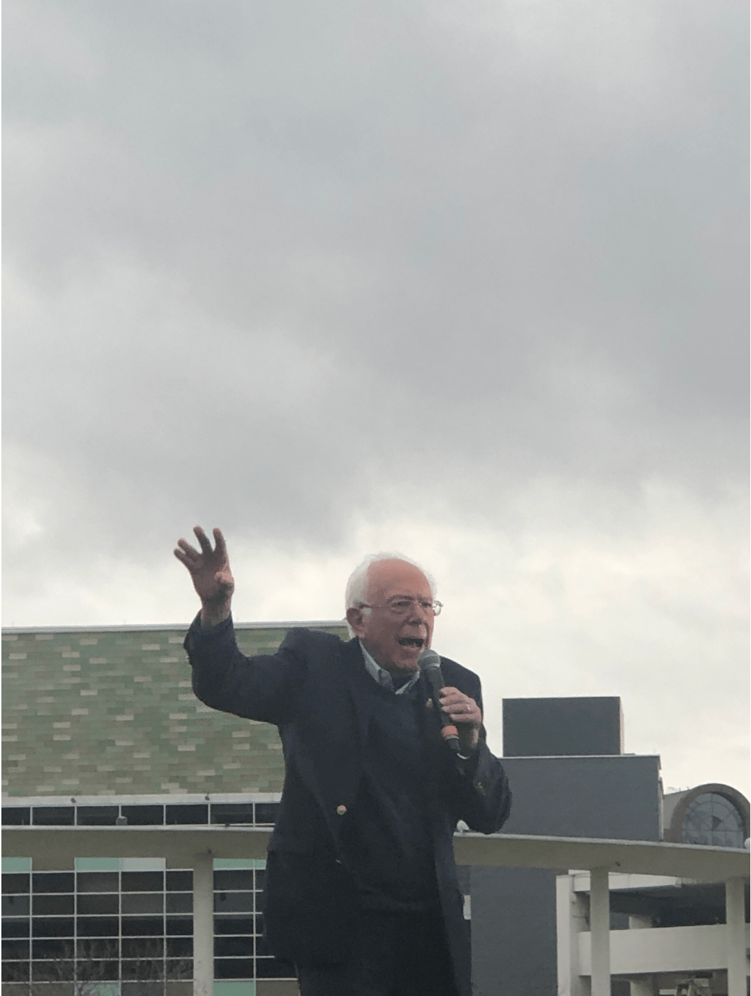 Bernie Sanders stands on stage surrounded by a crowd as he speaks into a microphone
