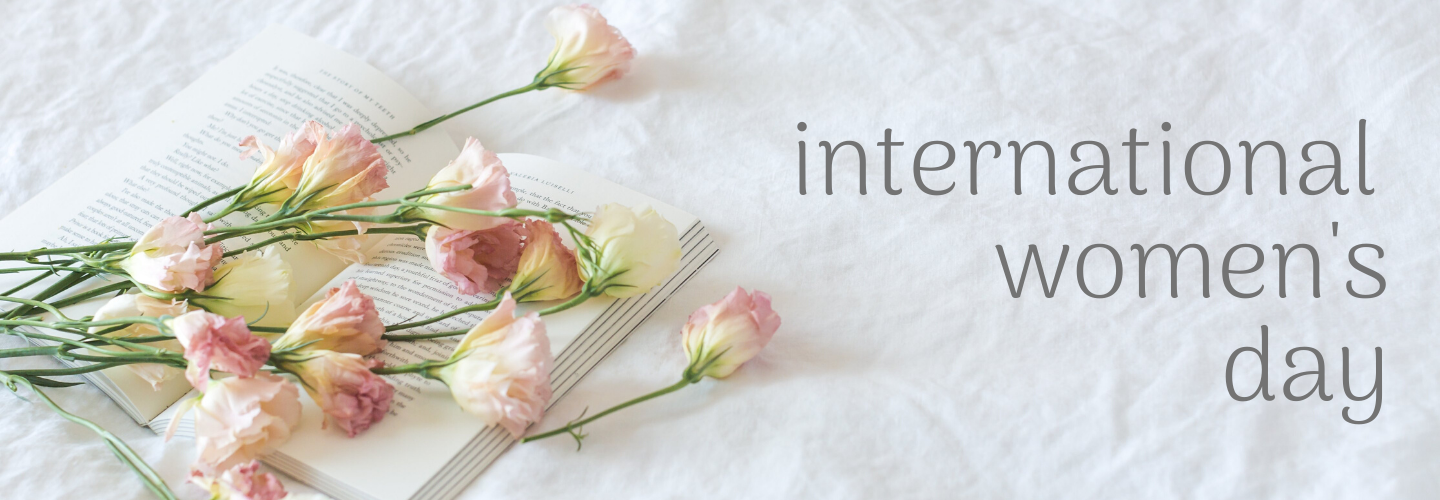 Light pink flowers laying across a book. Next to it, the words "international women's day" in a curvy, gray font.