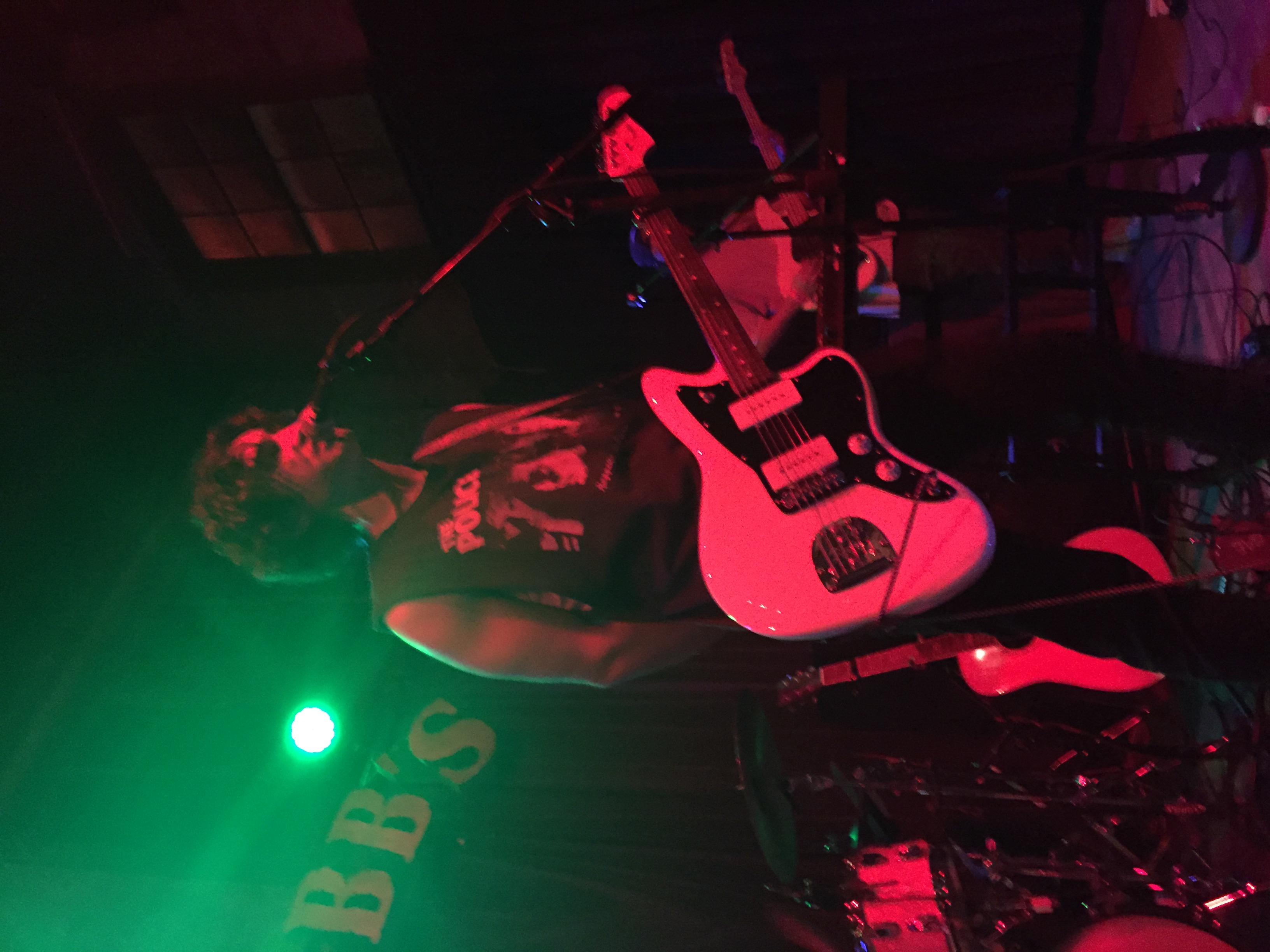 Graham is up on stage with a black and white electric guitar in his hands. He’s wearing all black as red tinted stage lights shining on him.